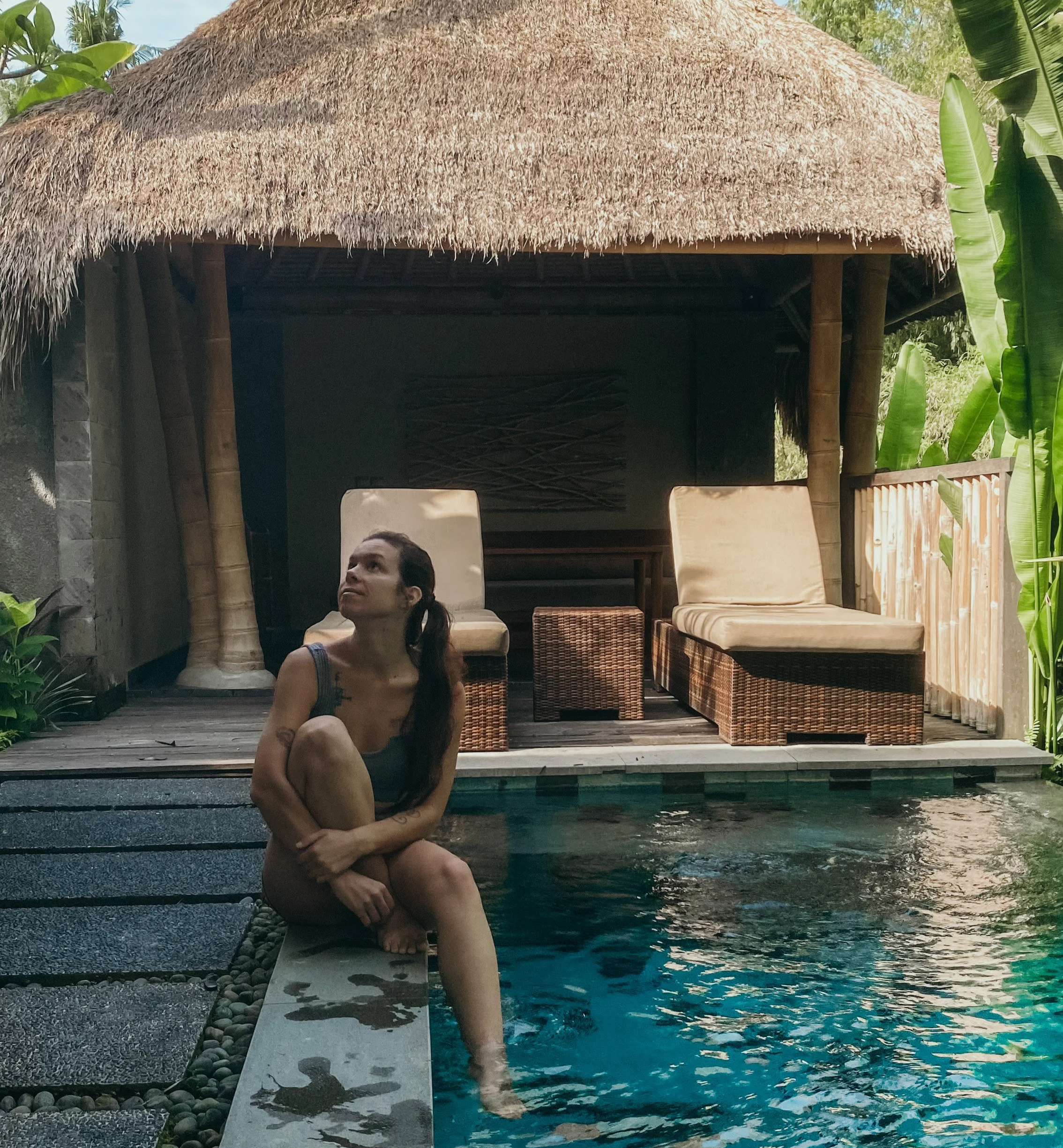 A woman sitting on the edge of a pool with tropical thatched hut and lounge chairs in the background, surrounded by lush greenery.