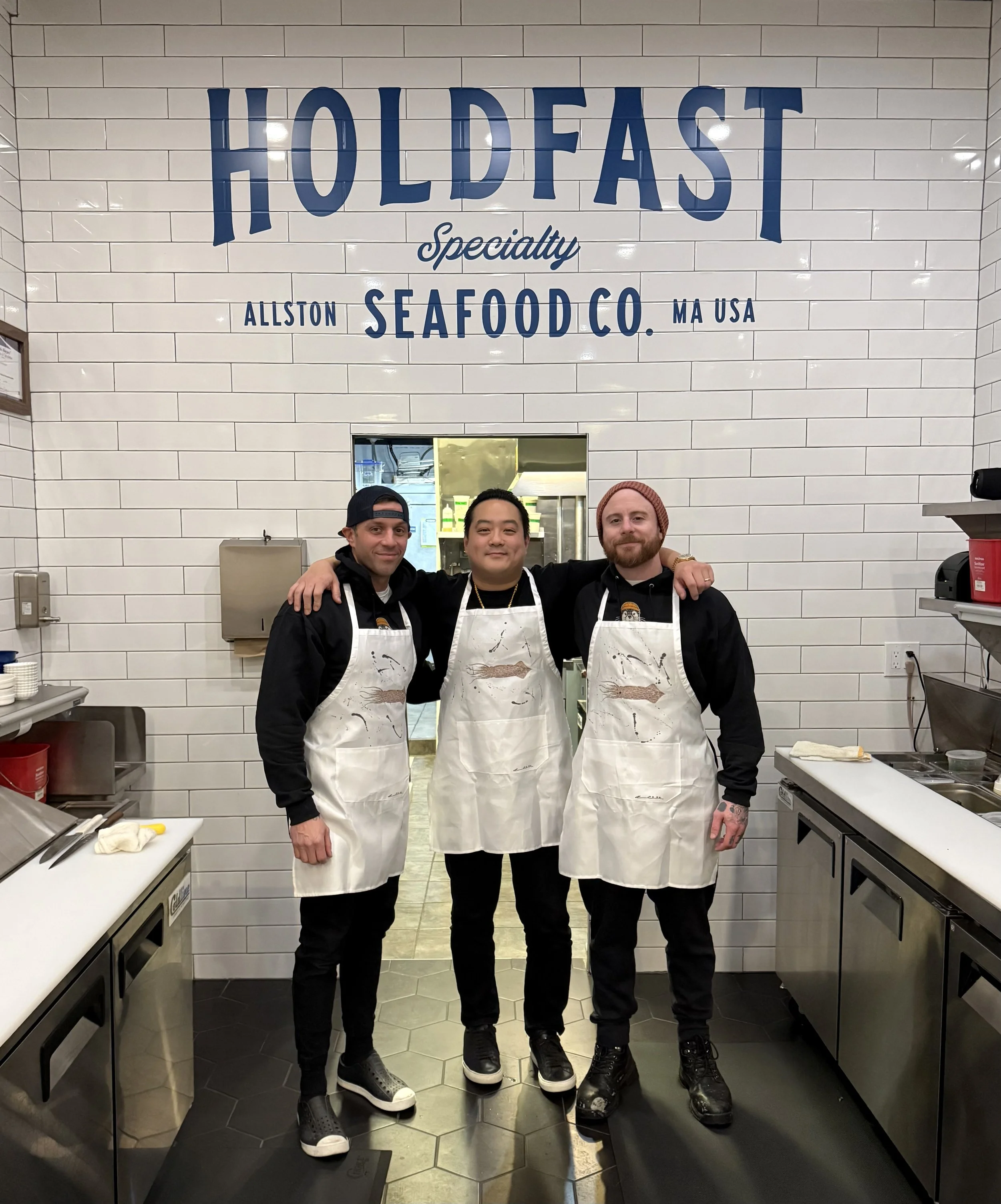 Three chefs in black uniforms and white aprons standing together inside a seafood restaurant called HOLDFAST, with a white tiled wall and a kitchen opening behind them.