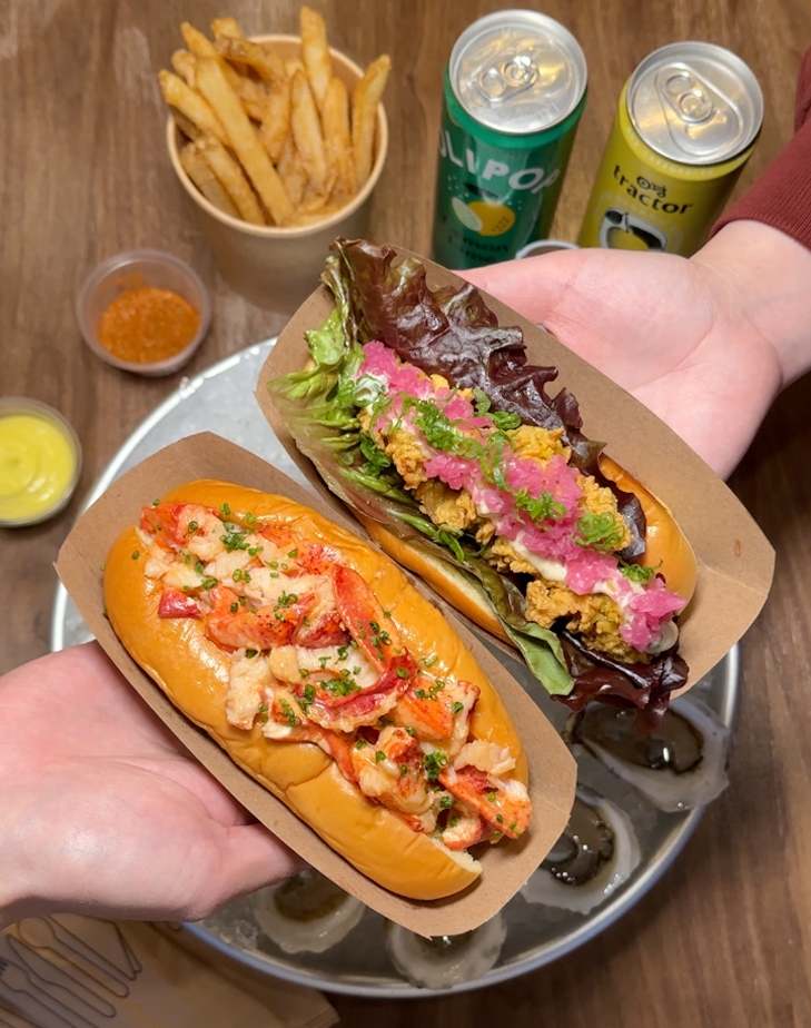 Close-up photo of two lobster rolls being held above a plate of raw oysters resting on a table. The table also has some soft drinks, dipping sauces and french fries.