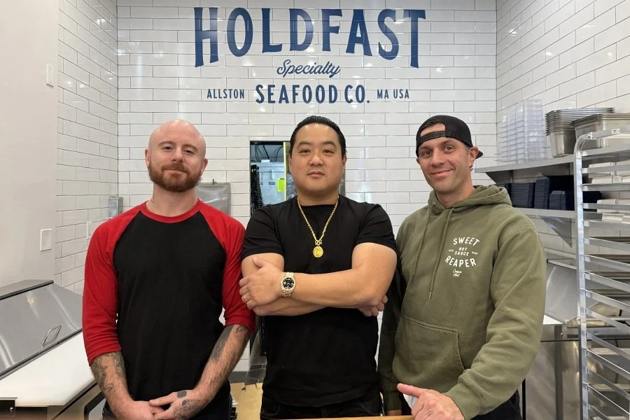 3 male subjects standing in an open air kitchen, in front of signage on tile