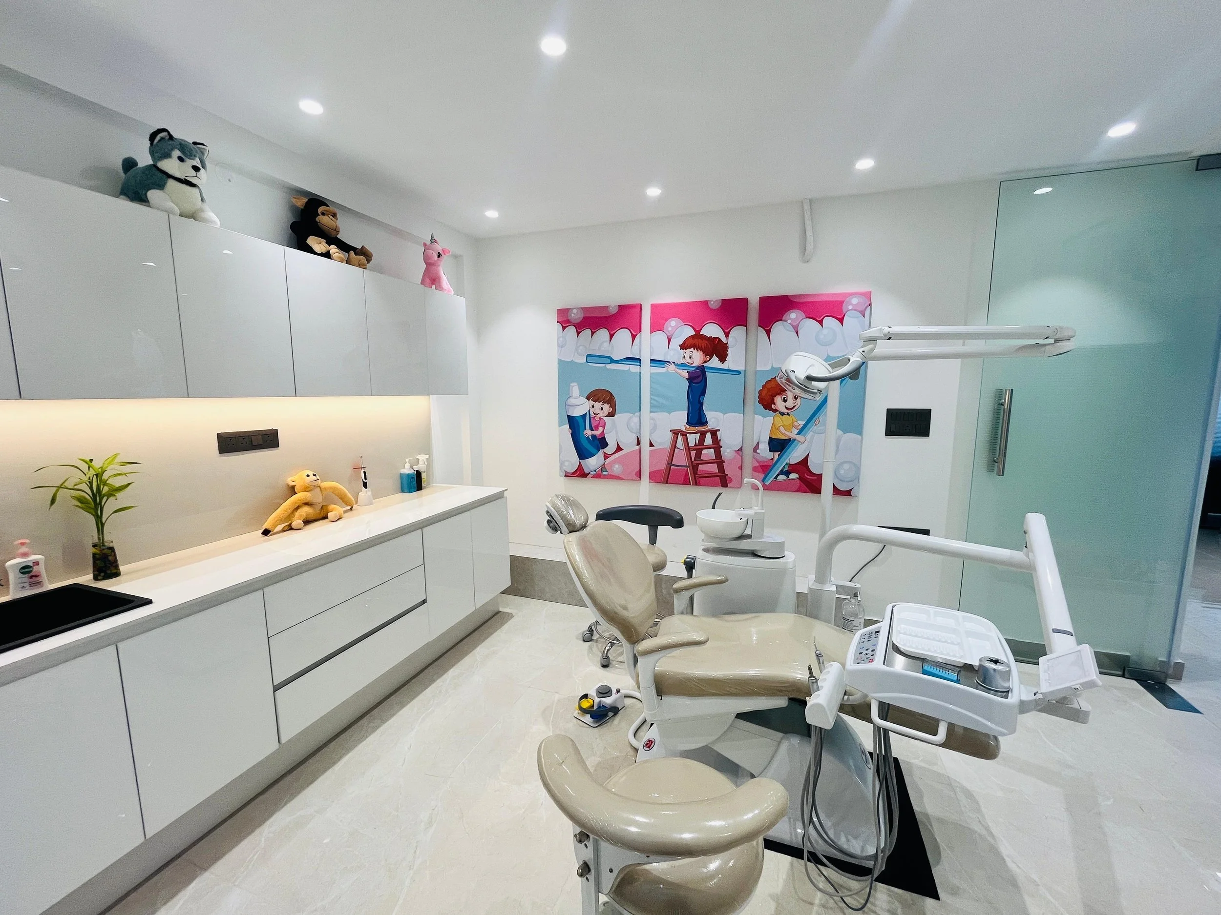 A dental treatment room with white cabinets, a beige dentist chair, and children's cartoon artwork on the wall. Plush toys are placed on top of the cabinets, and dental tools are on the chair's tray. A small plant and bottles are on the counter.