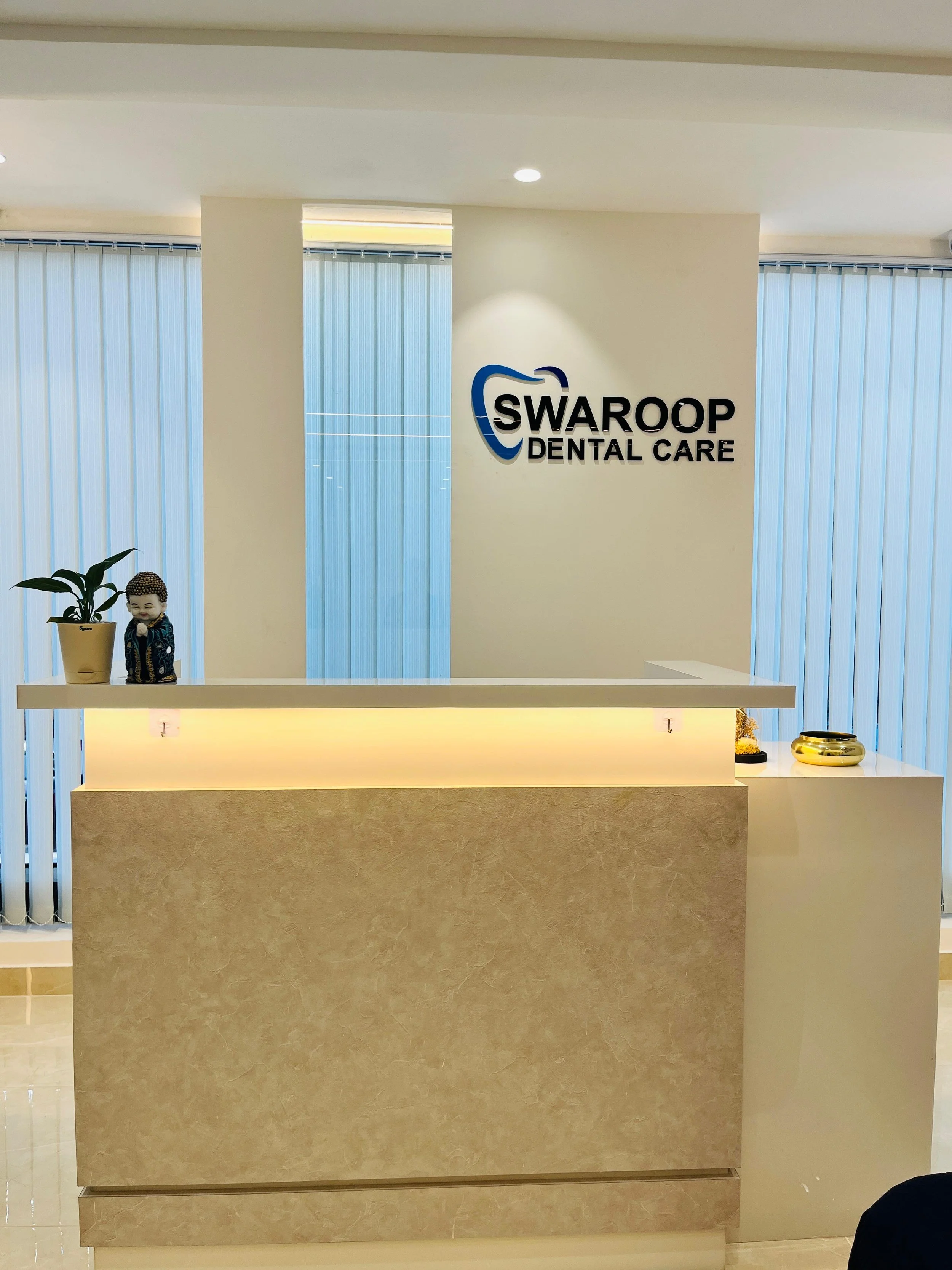 Reception desk at Swaroop Dental Care with a potted plant and decorative items, logo on the wall behind.