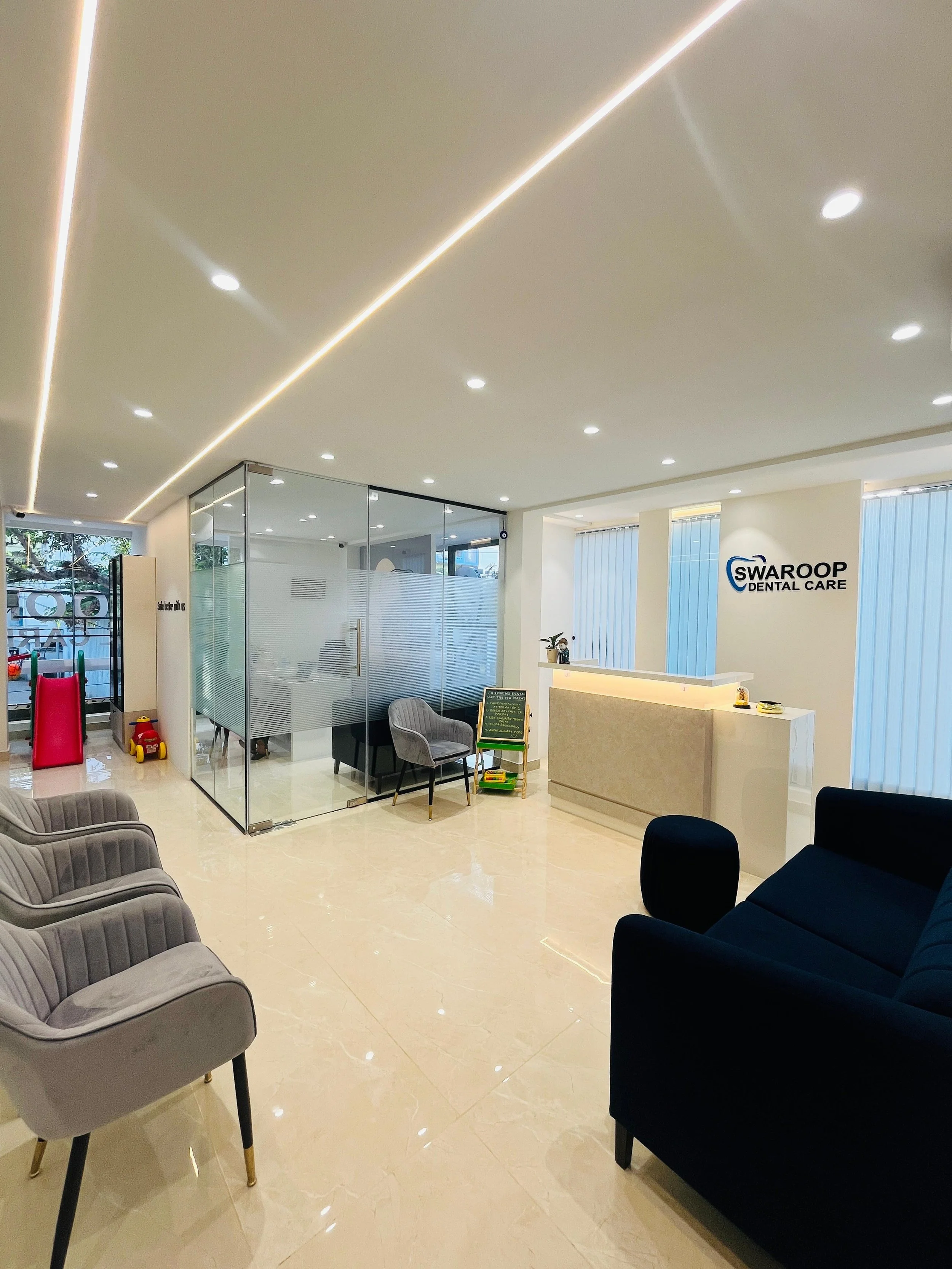 Modern dental office reception area with comfortable chairs, a glass-walled consultation room, and a front desk with a Swaroop Dental Care sign.