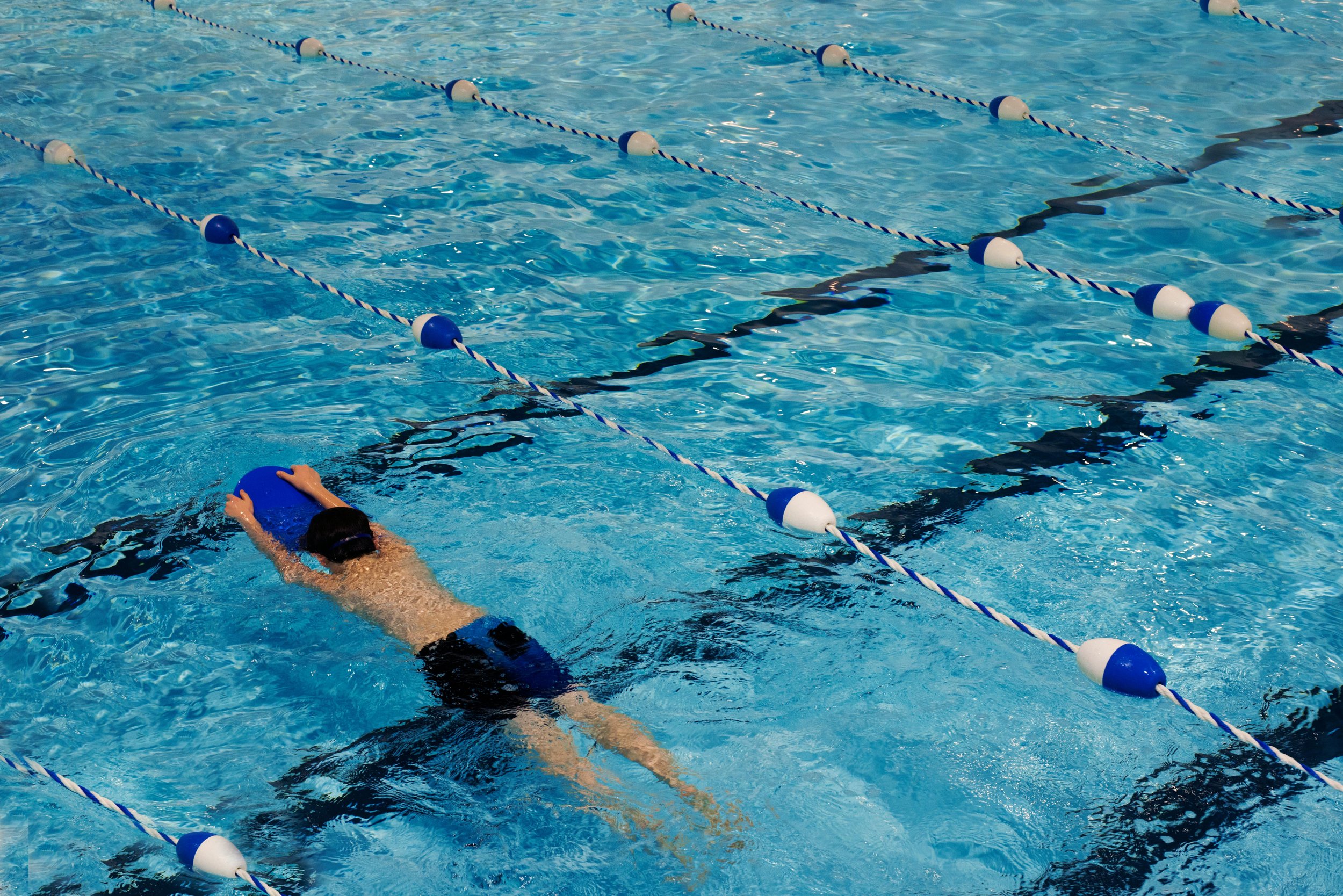 A person swimming in a pool, holding a blue kickboard, surrounded by lane dividers.
