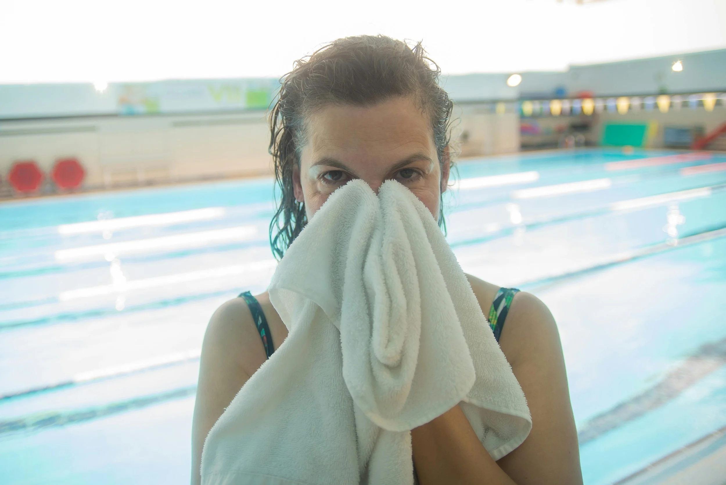 A woman wiping her face with a towel at an indoor swimming pool.