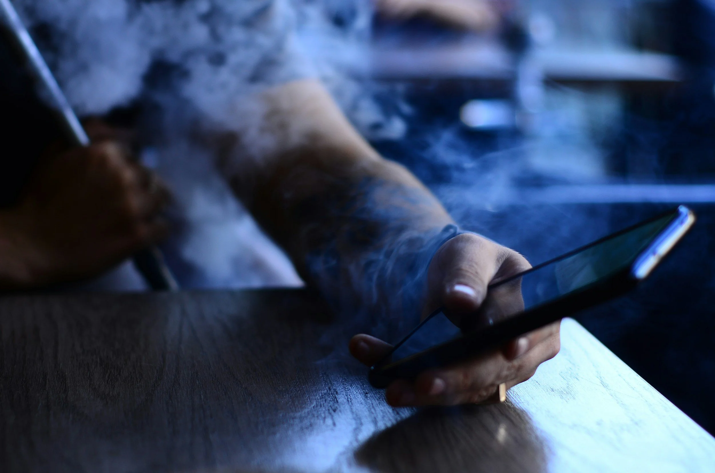 A person holding a smartphone with one hand and using a stylus with the other, sitting at a wooden table with smoke around their arm.