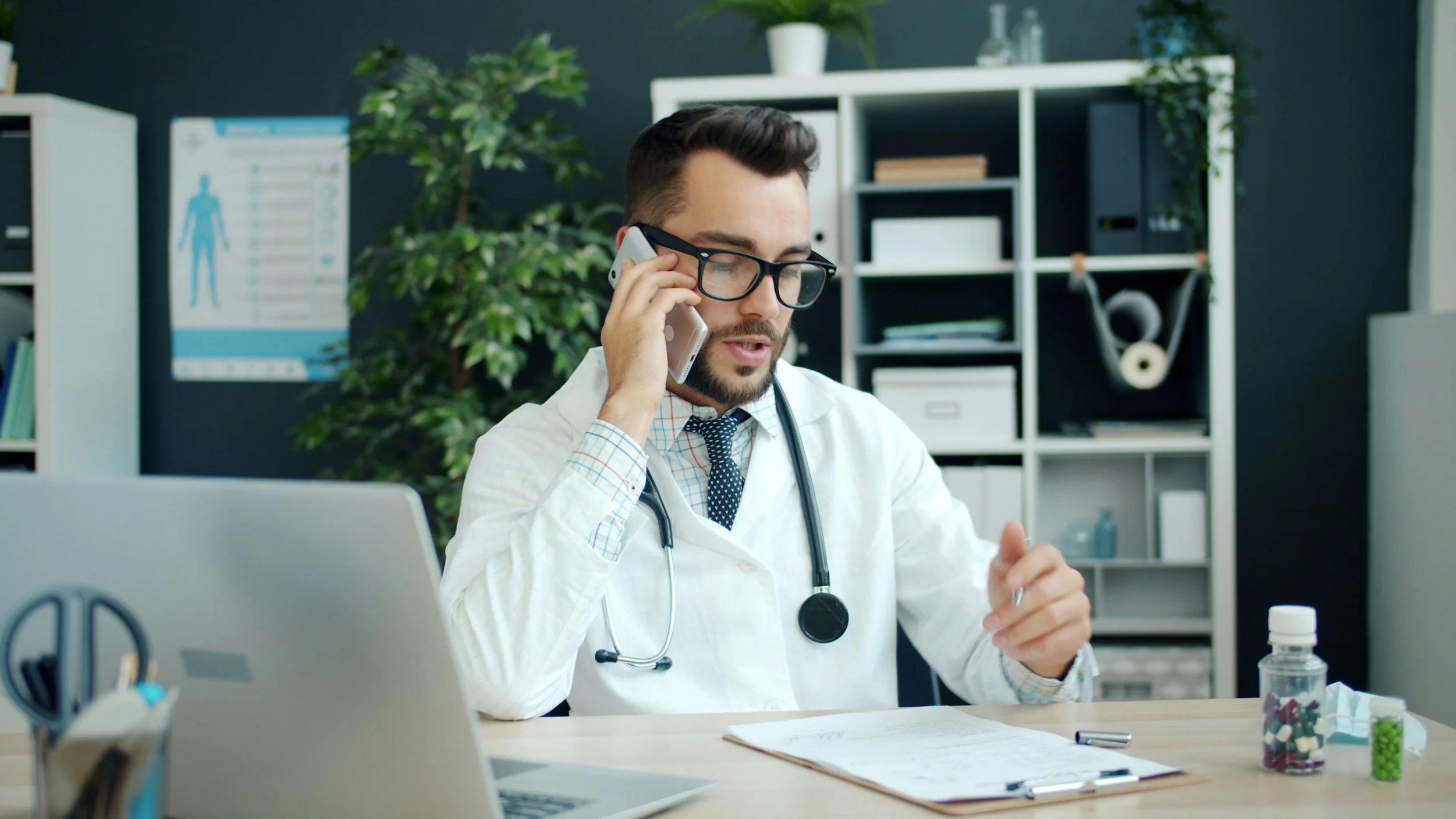 A male doctor wearing glasses and a white coat with a stethoscope around his neck, talking on the phone at his desk in a medical office.