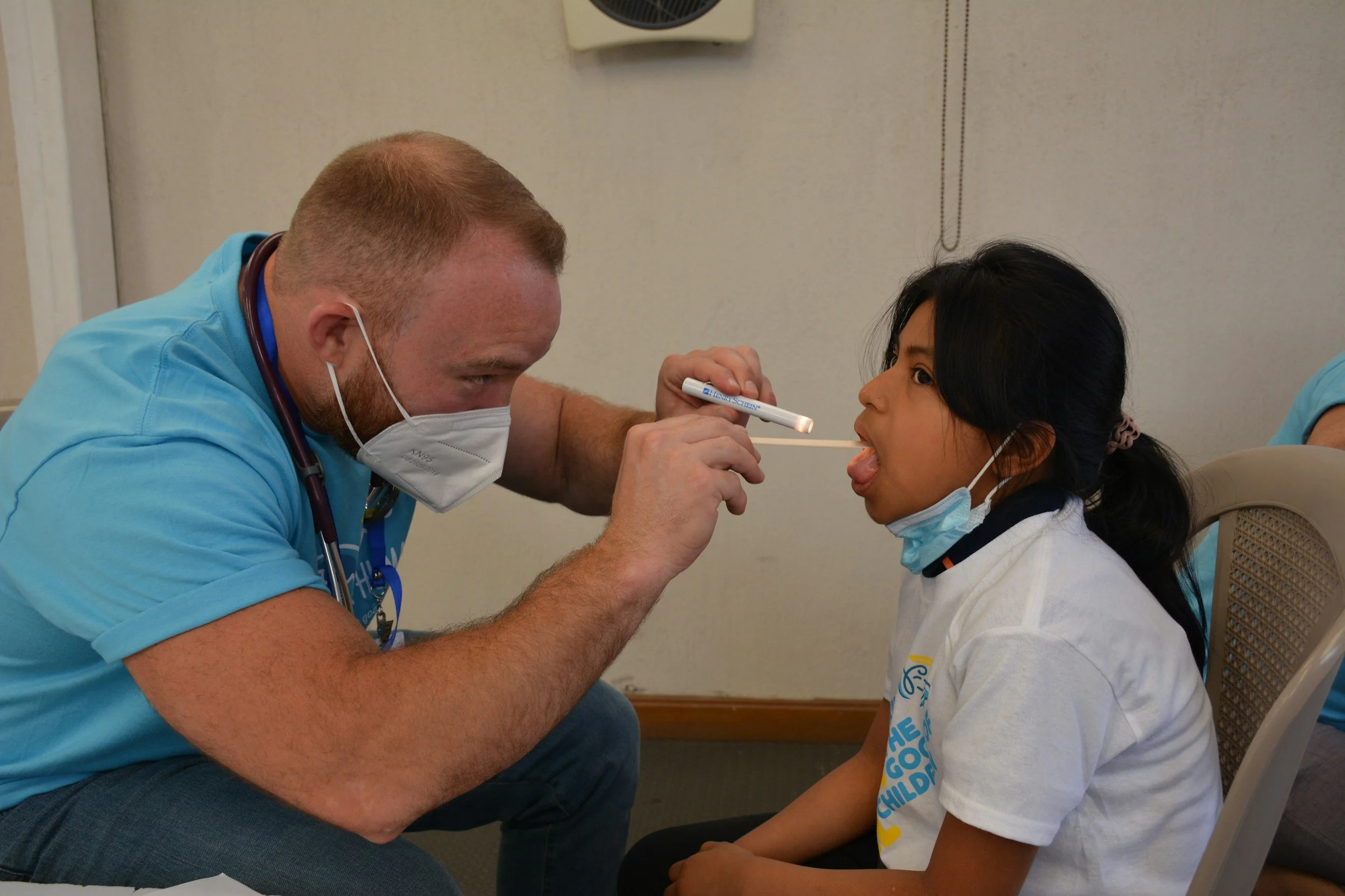 A healthcare worker administers a COVID-19 test to a young girl by inserting a nasal swab into her nostril. The girl is sitting on a chair, wearing a white T-shirt and face mask pulled down under her chin. The healthcare worker wears a blue uniform, a face mask, and has a stethoscope around his neck.