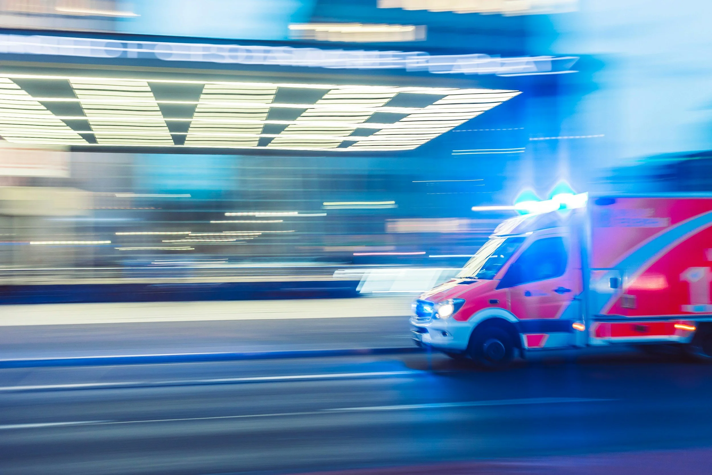 Blurred motion image of a red emergency vehicle with flashing lights driving past a modern building.