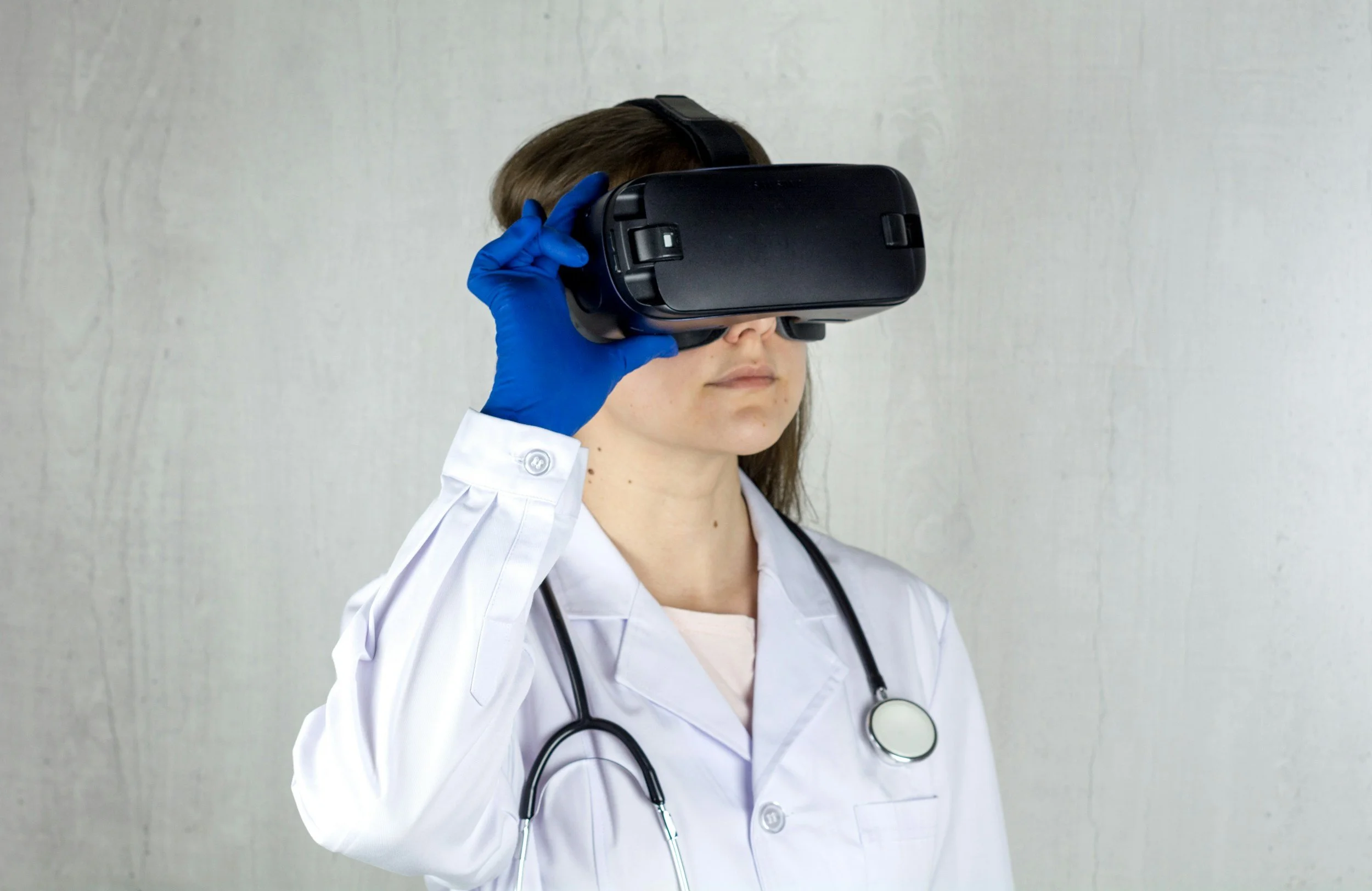 A female medical professional wearing a white coat, blue gloves, and a stethoscope around her neck, looking through a virtual reality headset.