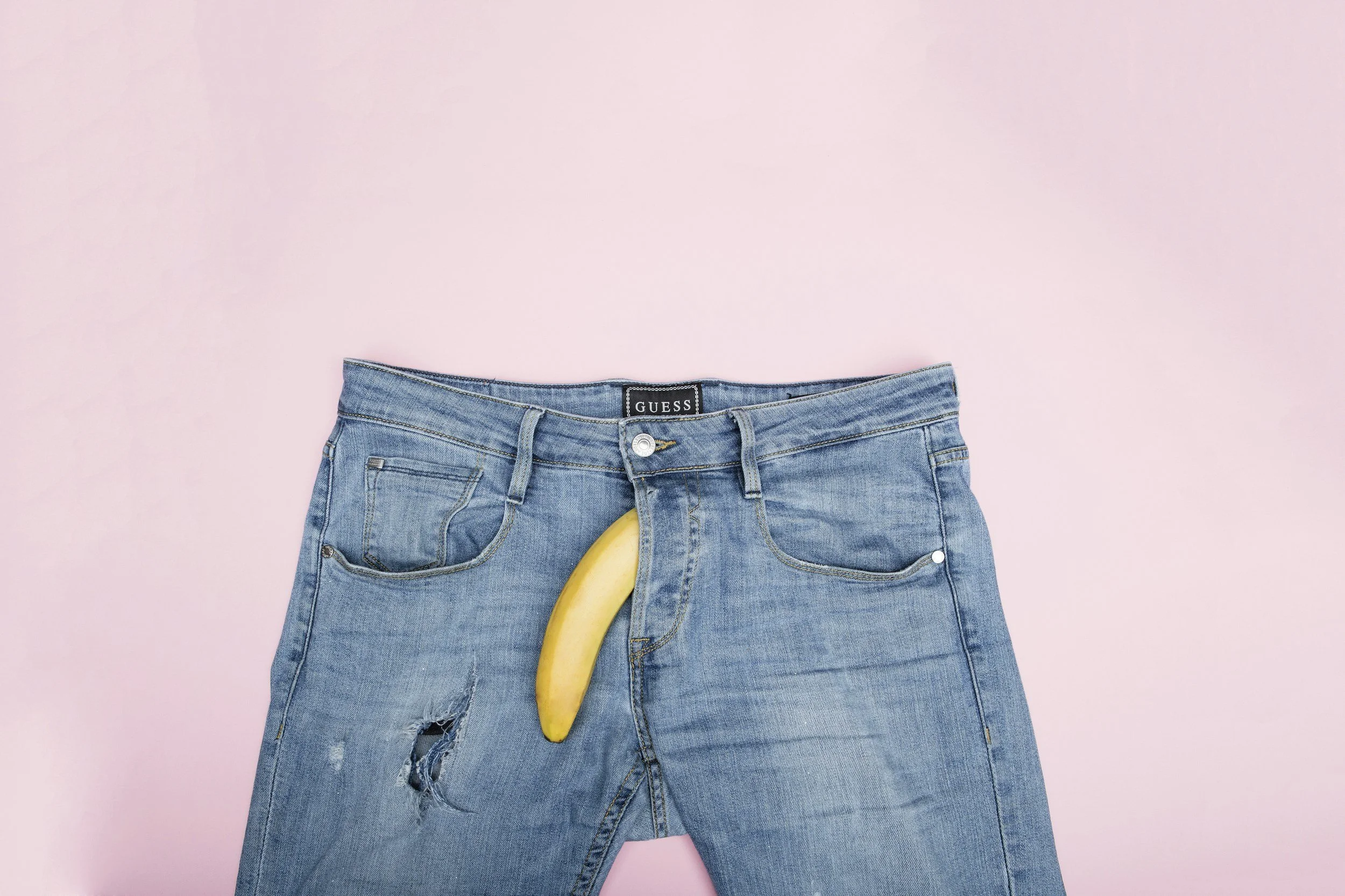 A pair of blue jeans on a pink background with a banana placed in the front pocket.