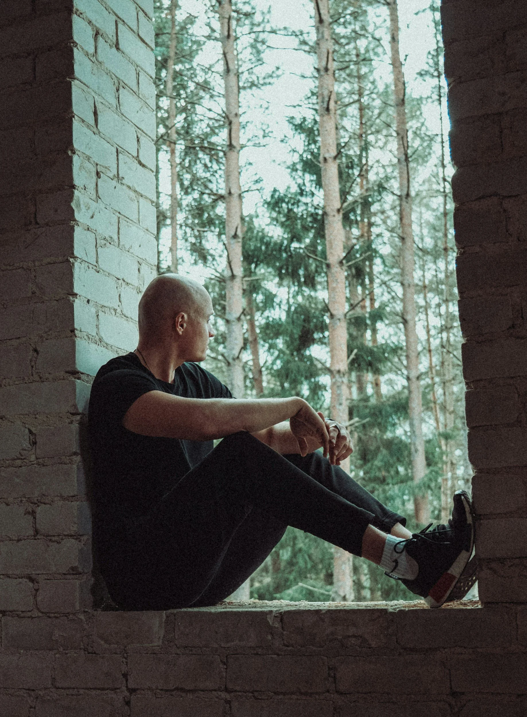 A person with a shaved head sitting on the ground inside a brick structure, looking out at tall trees in a forest.