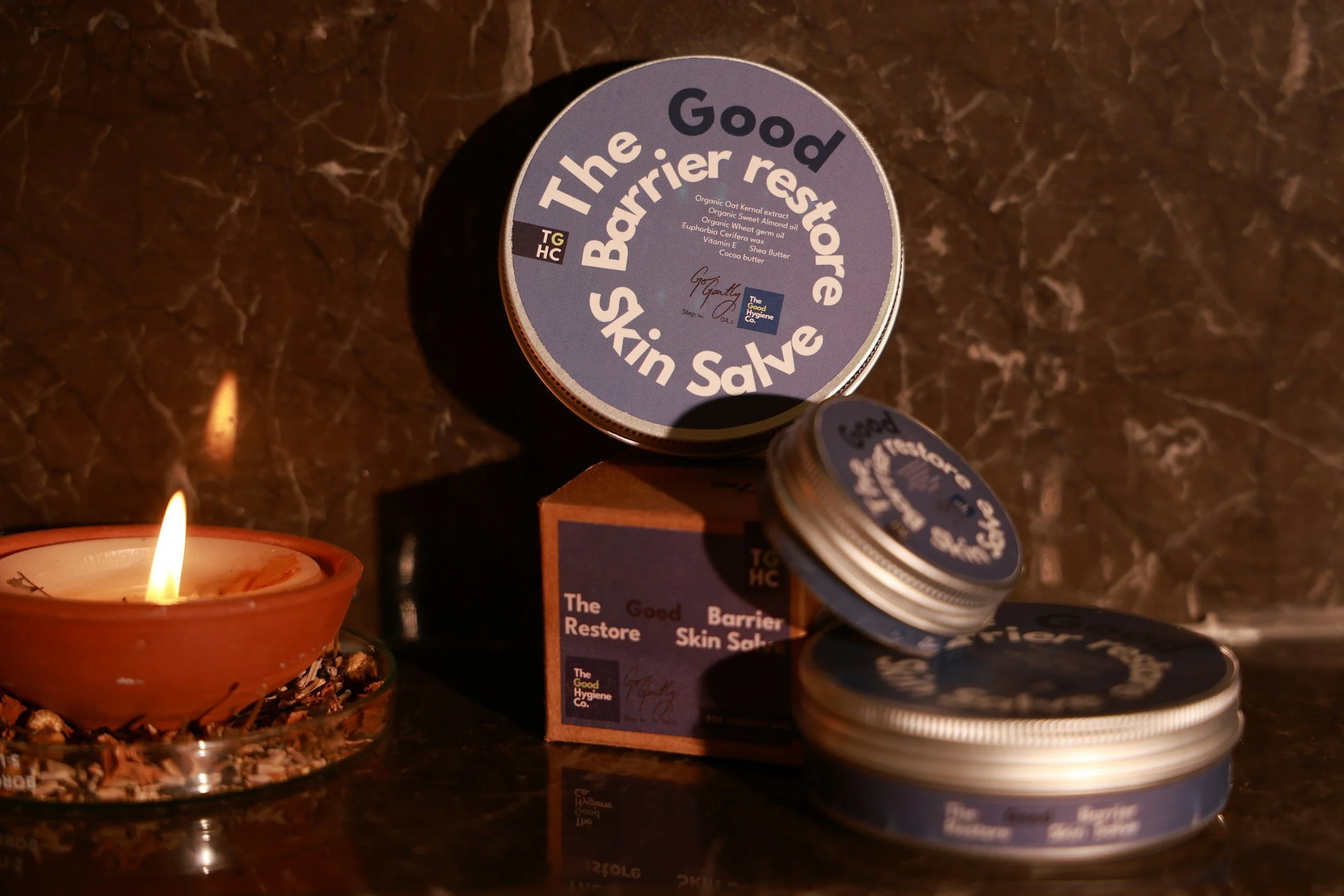 A round container labeled 'The Good Barrier Restoring Skin Save' with ingredient list, placed on a wooden block, surrounded by smaller tins and a lit candle in an orange holder on a dark, reflective surface.