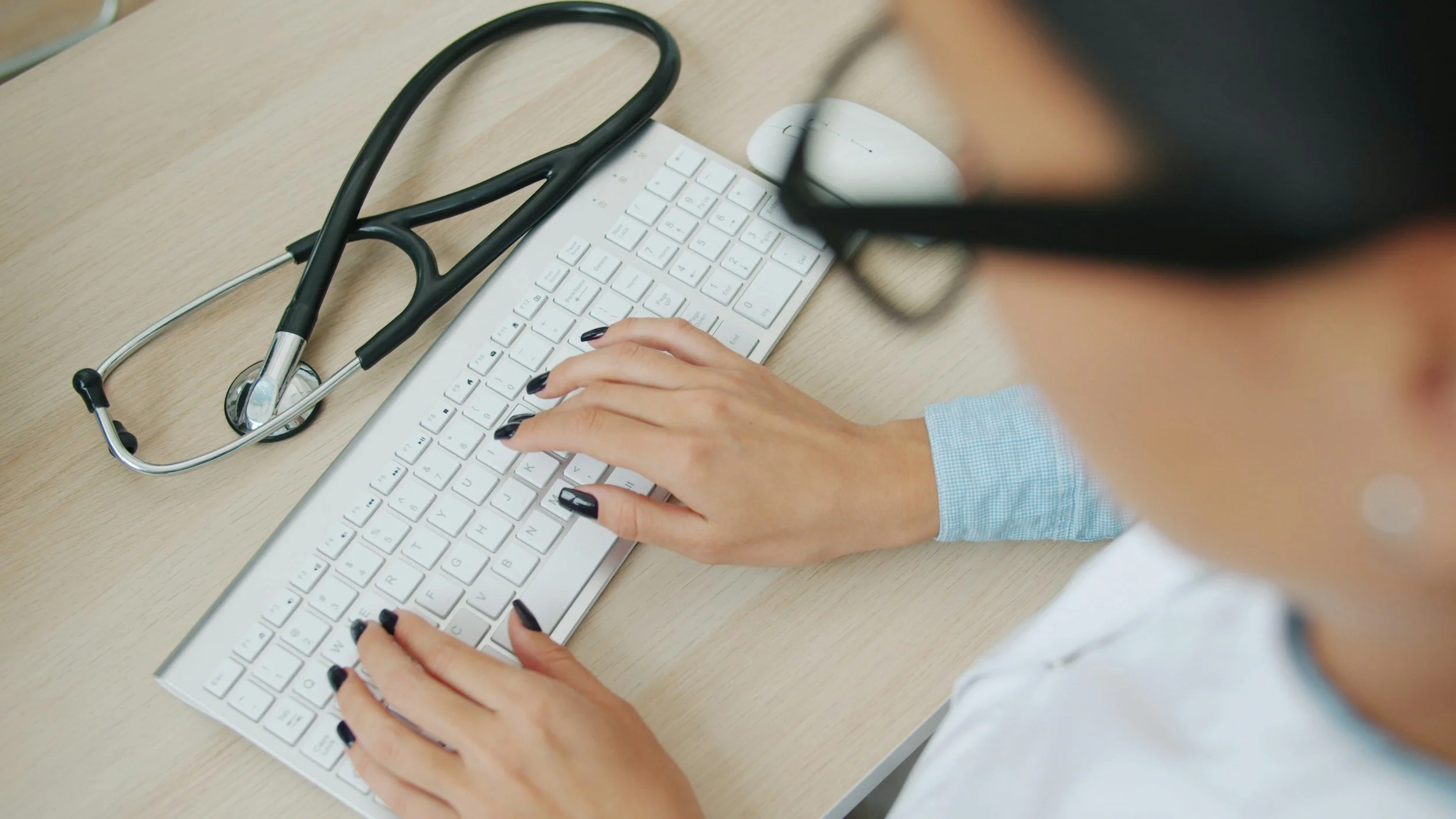 A person with black painted fingernails is typing on a white keyboard at a wooden desk, with a stethoscope placed on the keyboard.
