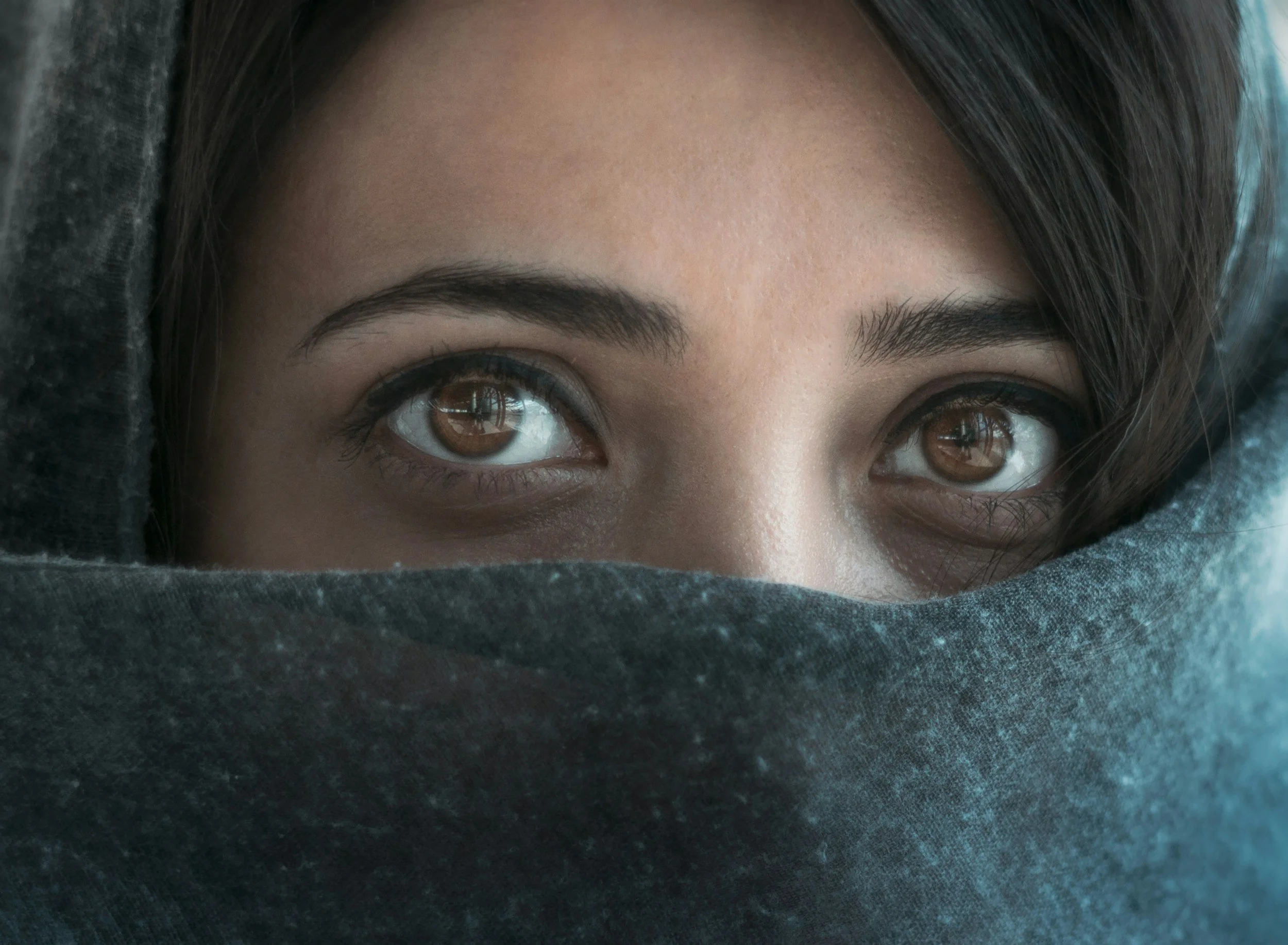 Close-up of a woman's face with brown eyes and dark eyebrows, partially covered by a dark hoodie.