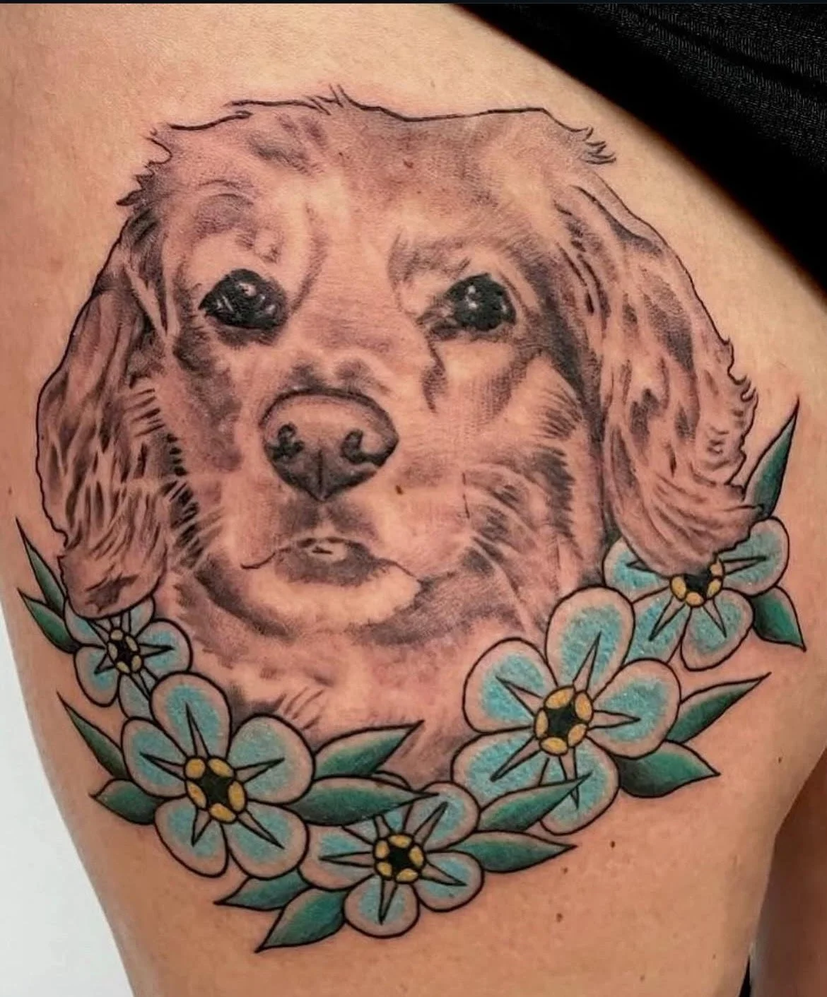 A lifelike black and grey dog portrait surrounded by bright blue flowers. The combination of realism and traditional linework creates a heartfelt memorial feel.