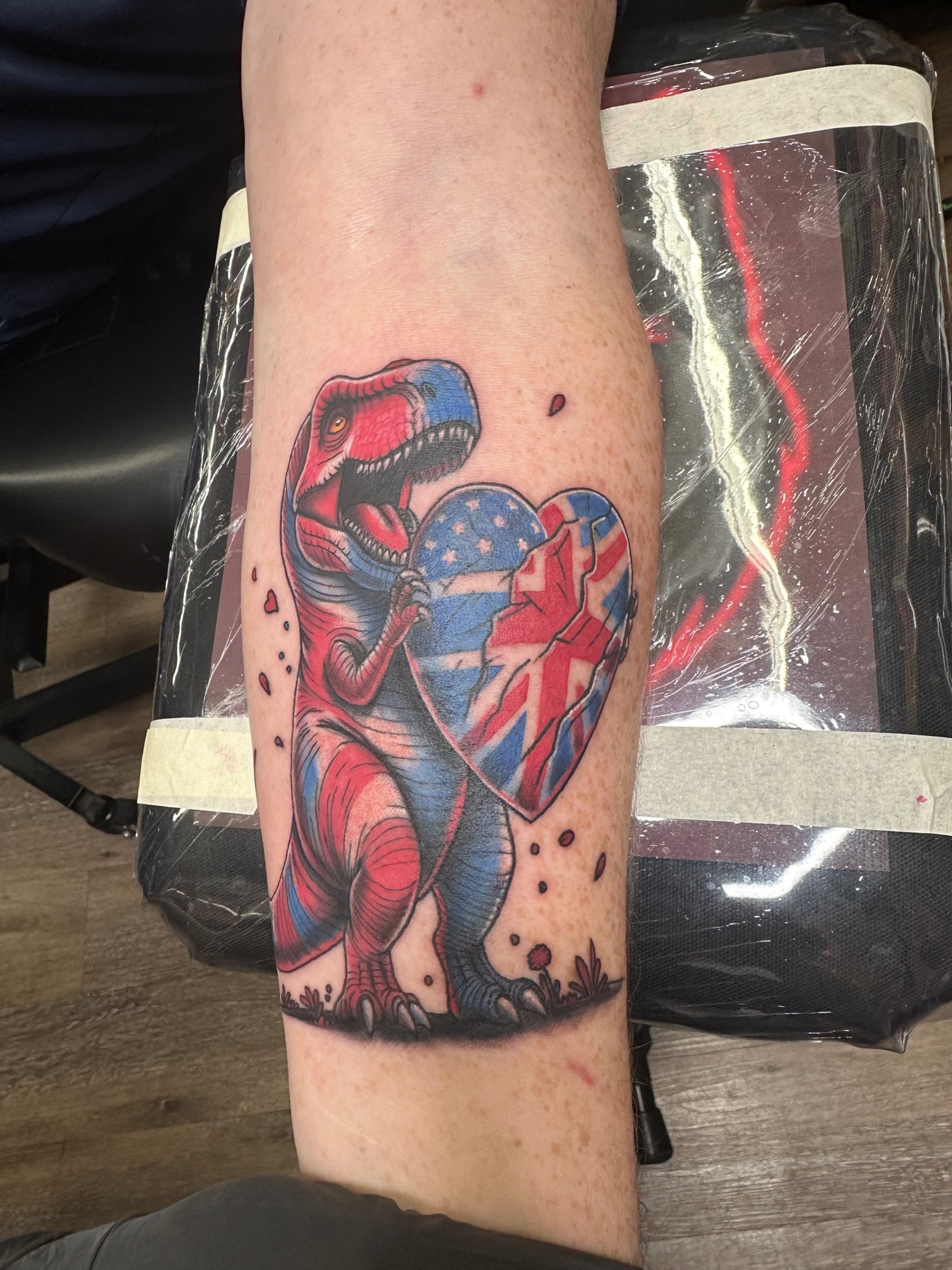 A detailed red, white, and blue T-Rex clutching a heart patterned with American and British flags. The sharp linework and saturated colors make this piece fierce, fun, and eye-catching.
