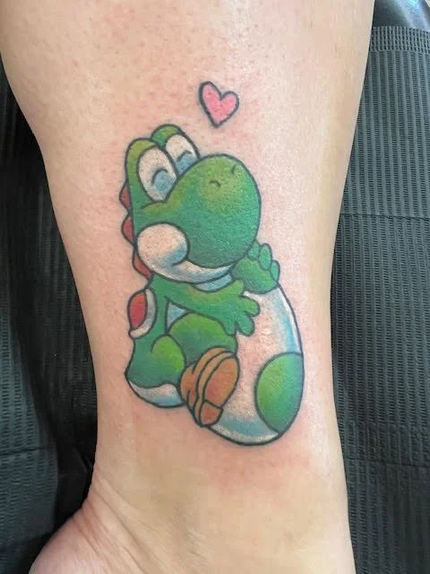 A bright, playful tattoo of Yoshi from the Mario universe, hugging his egg with a heart overhead. The vibrant color work and soft shading bring this nostalgic character to life with warmth and personality.