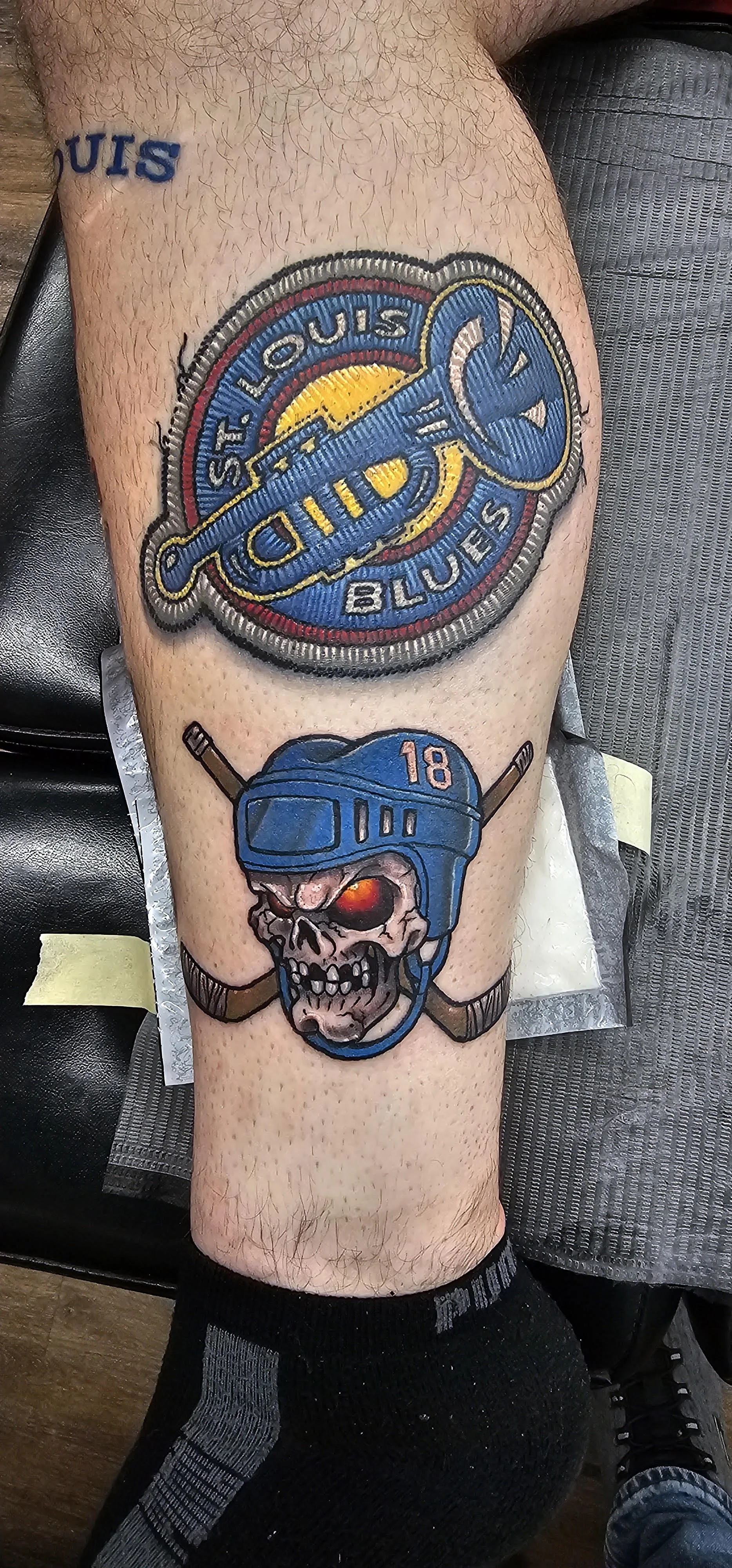 This tattoo mimics the look of an embroidered patch for the St. Louis Blues logo, complete with textured stitch detailing and layered color shading. Below it, a fiery-eyed skull in a hockey helmet adds a dramatic, gritty edge with comic-book energy.