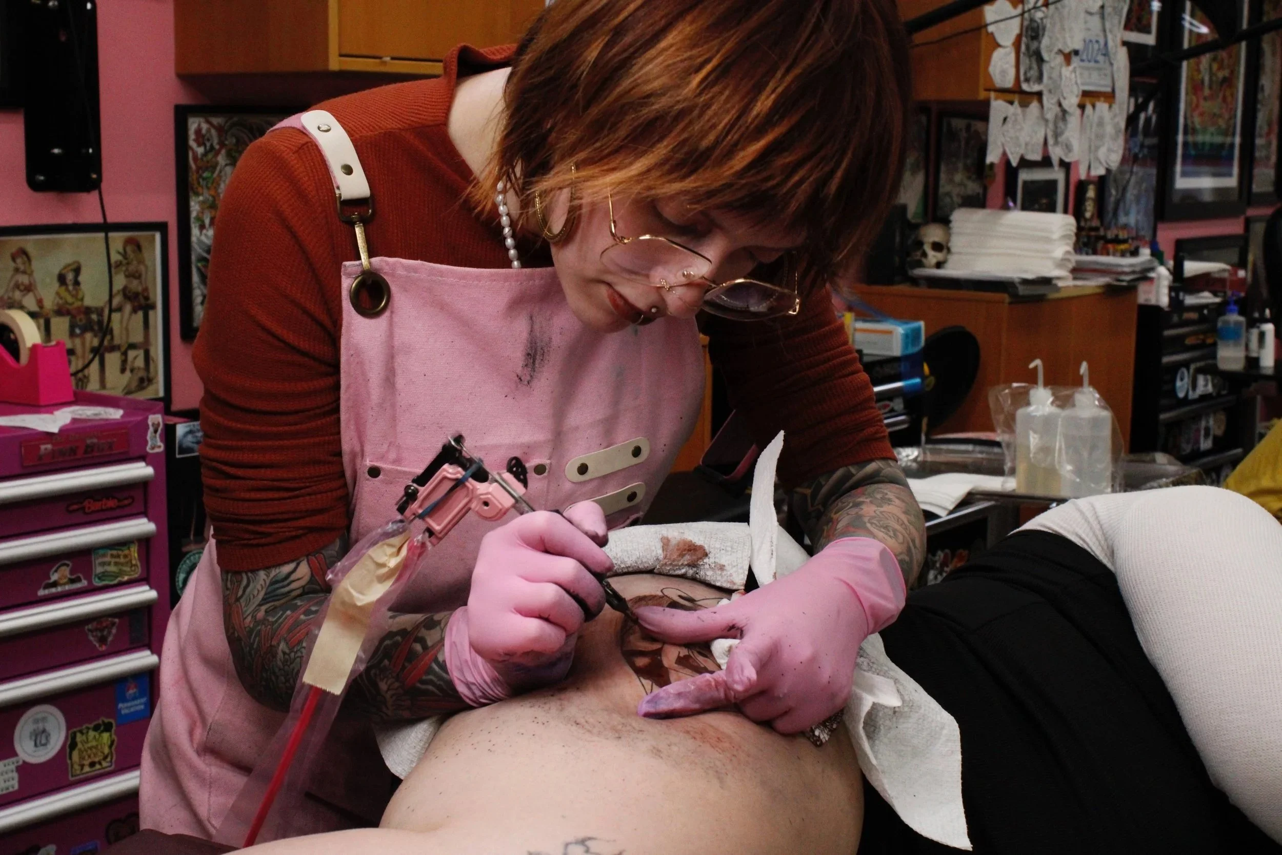 Tattoo artist wearing pink gloves and glasses working on a person's tattoo in a tattoo studio, surrounded by tattoo supplies and artwork on the walls.