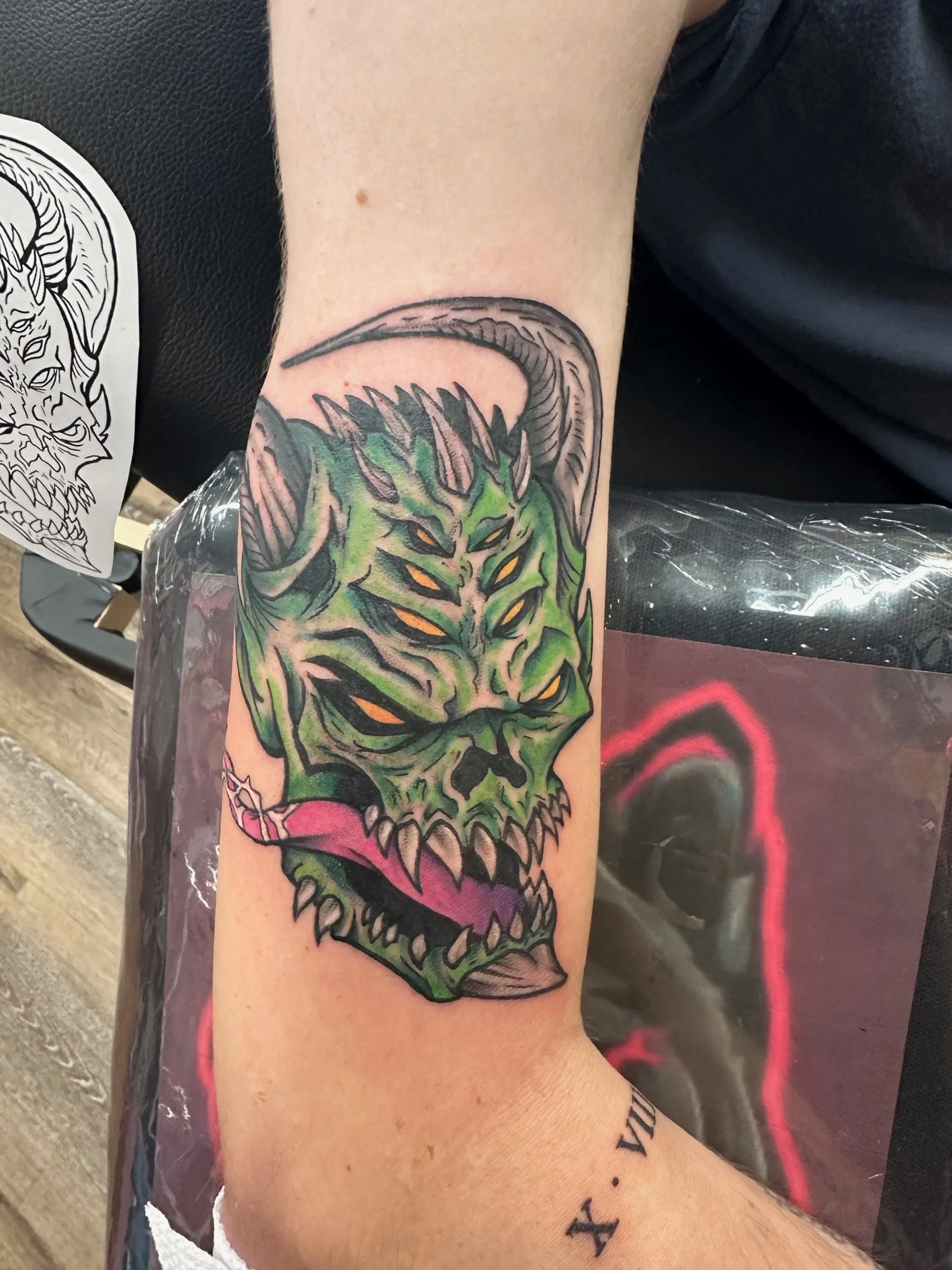 A vivid, neo-traditional depiction of a snarling green demon with intense yellow eyes and sharp, exaggerated features. The dynamic color blending and bold outlines give it a fierce, high-energy presence.