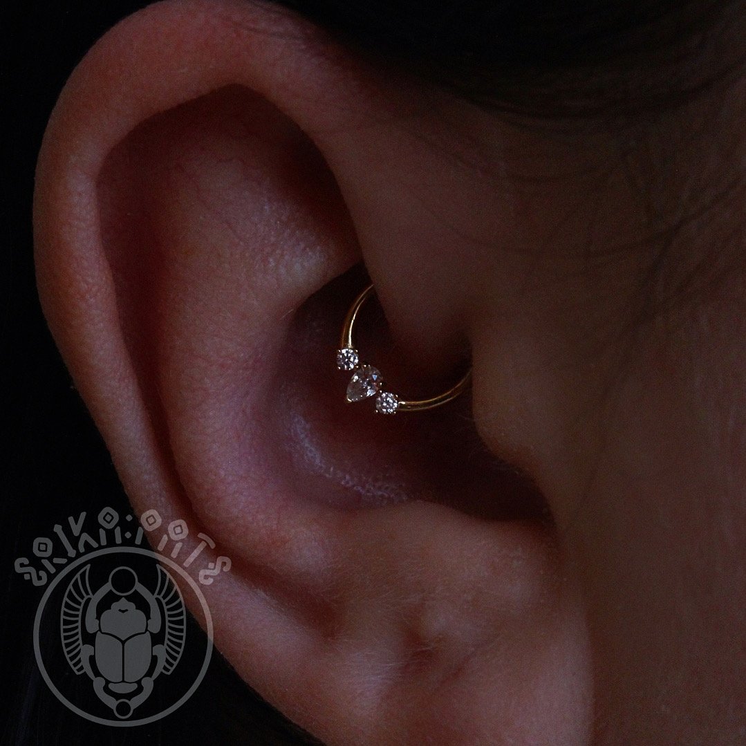This daith piercing features a gold ring tucked neatly into the inner fold of the ear’s cartilage. Its placement adds depth and dimension to any curated ear while offering a unique, understated look.