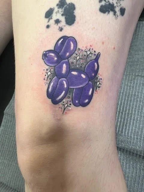 A shiny purple balloon dog rendered with reflective highlights and dotted background. The vibrant coloring and playful tone make this a modern pop-art style tattoo.