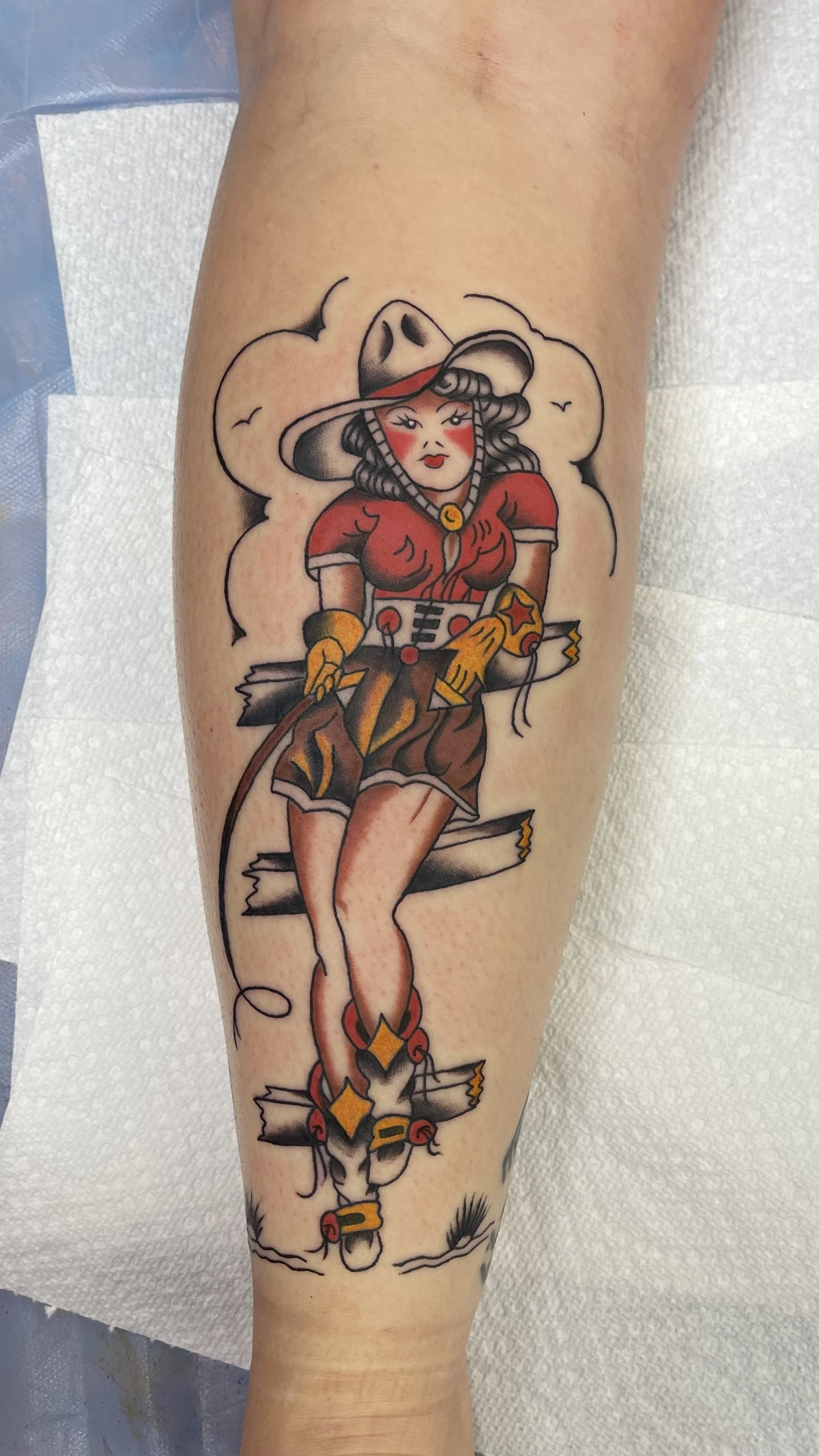 This bold traditional-style tattoo features a confident cowgirl posed against a rustic wooden fence, complete with a hat, whip, and western flair. The saturated reds, yellows, and clean black lines give it that classic Americana charm and vintage pin
