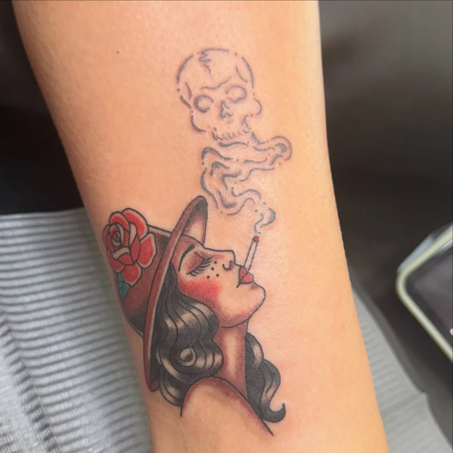 A neo-traditional style tattoo featuring a woman in a hat, cigarette smoke swirling into a skull above her. The bold lines, red tones, and vintage aesthetic create a dramatic yet timeless piece.
