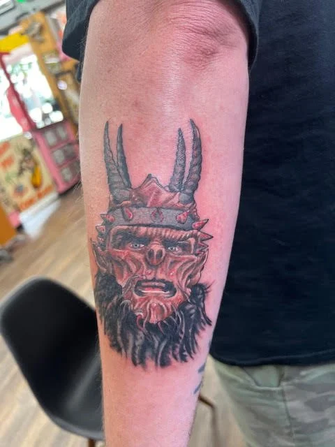A red-skinned horned warrior head with sharp details and rugged expression. The bold use of red and black ink gives this tattoo a powerful, mythic quality.