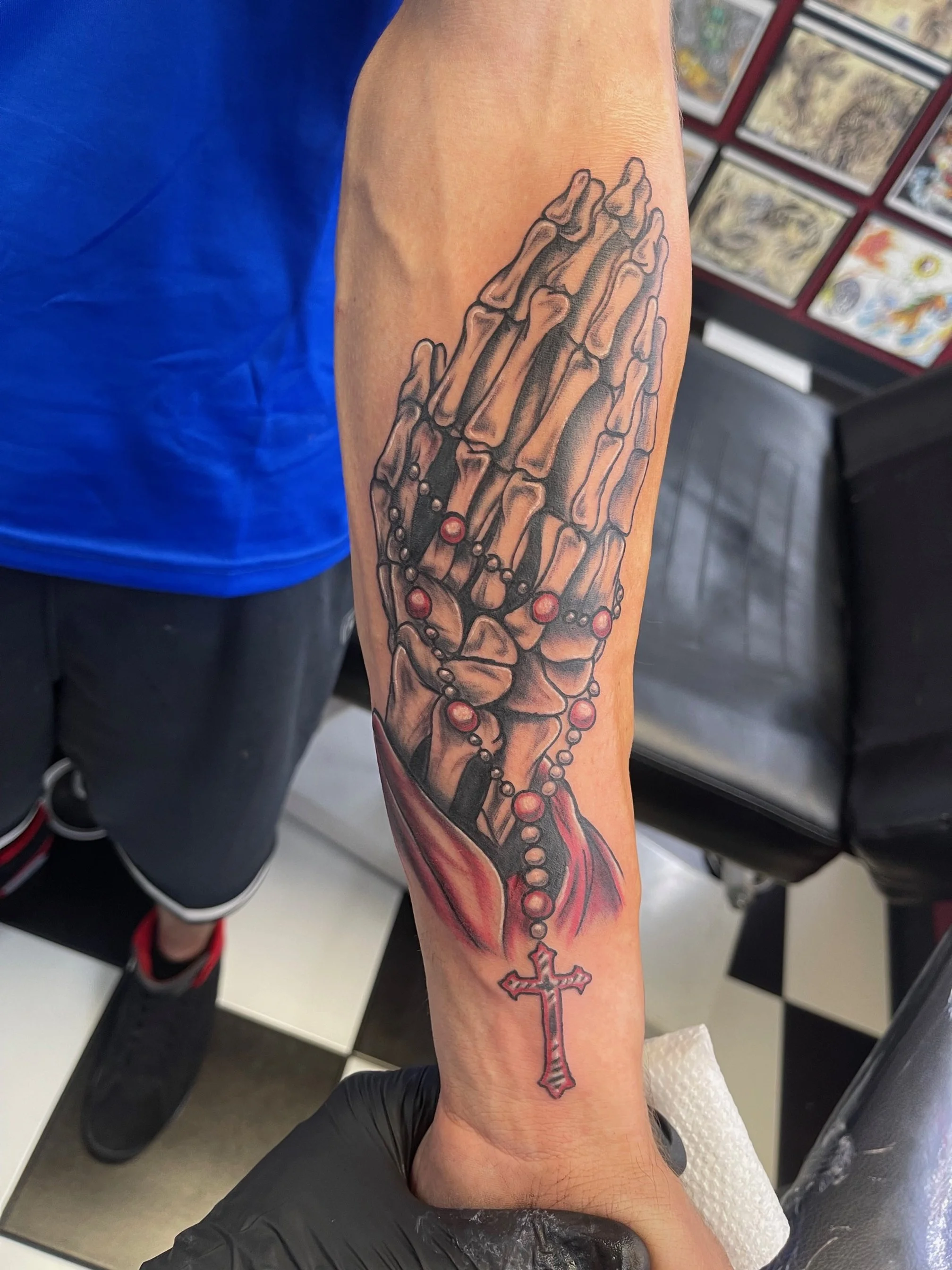 A realistic skeletal hand holding a rosary, with deep shading and subtle red highlights. The blend of anatomy and symbolism gives this design both spiritual and gothic undertones.