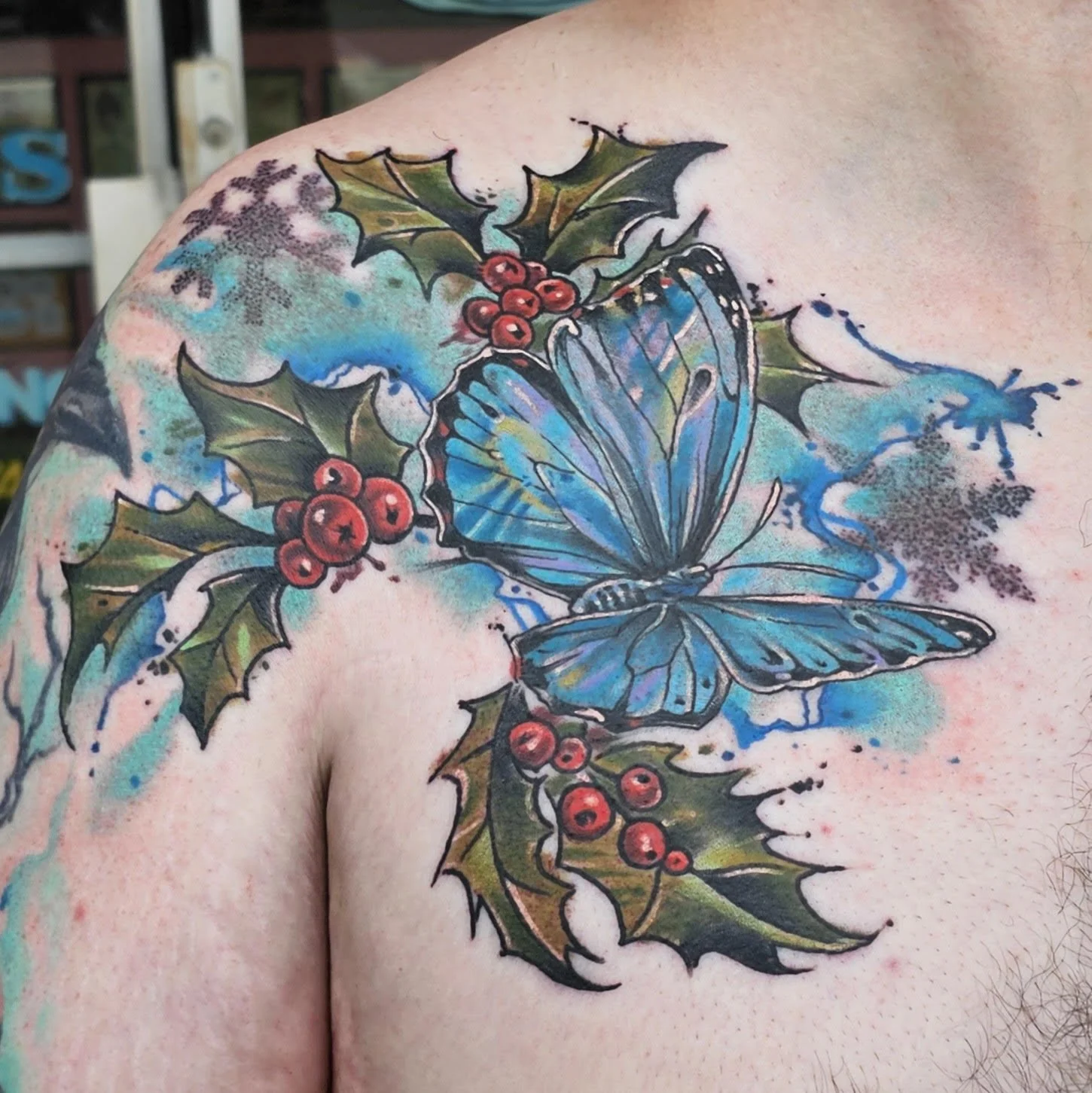 A detailed blue butterfly spreads its wings over a cluster of holly leaves and berries, set against a splashy watercolor background. The contrast of crisp linework and soft ice-toned textures gives it a wintery, ethereal feel.