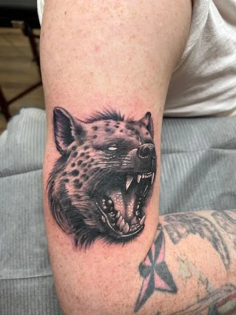 A realistic black and grey depiction of a snarling hyena, showcasing fine texture and lifelike expression. The attention to shading and depth gives this piece a fierce, powerful presence.