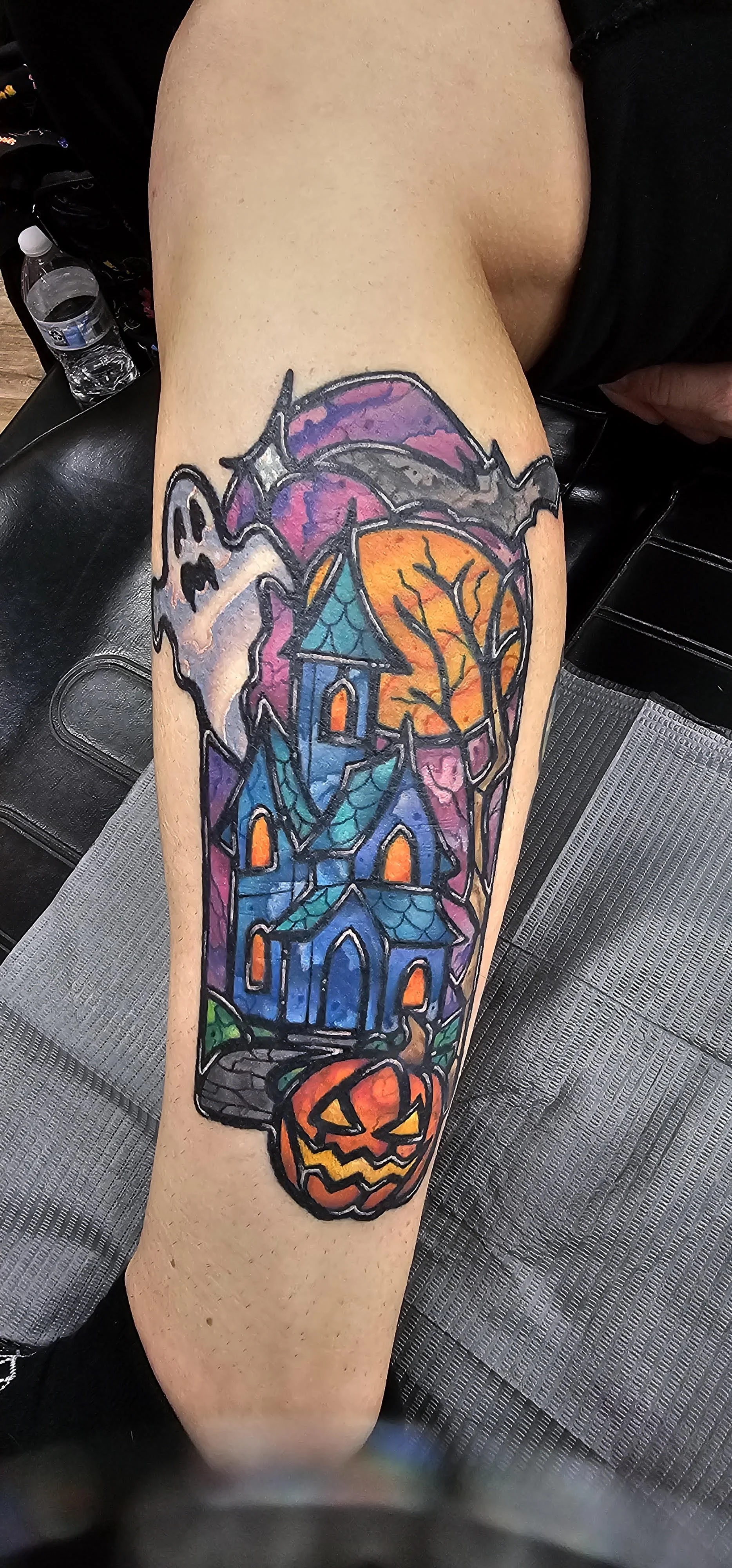 A stylized haunted house rises from a vivid, cartoon-inspired Halloween scene with a glowing moon, ghost, and grinning pumpkin. Bold outlines and saturated purples, blues, and oranges give it a fun yet spooky stained-glass vibe.
