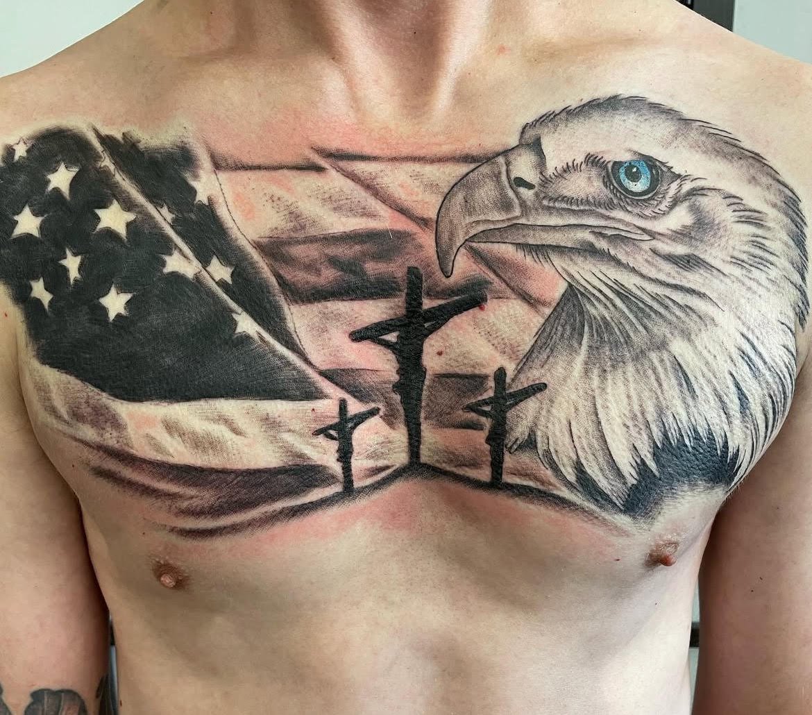 A large chest tattoo featuring a detailed eagle beside a waving American flag and three crosses. The imagery powerfully blends symbolism of strength, faith, and freedom.