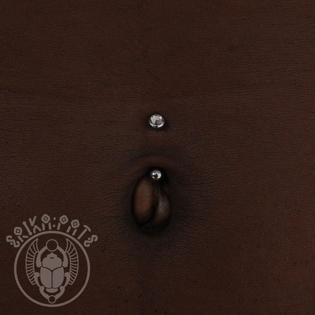Close-up of a person's navel piercing with two silver balls on a dark skin background. There is a logo in the bottom left corner with a beetle design and text.