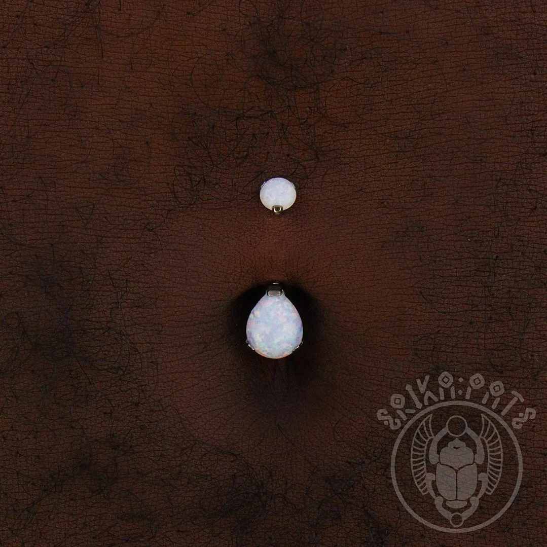 Close-up of a belly button with a piercing featuring a white bead and a teardrop-shaped opal gemstone.