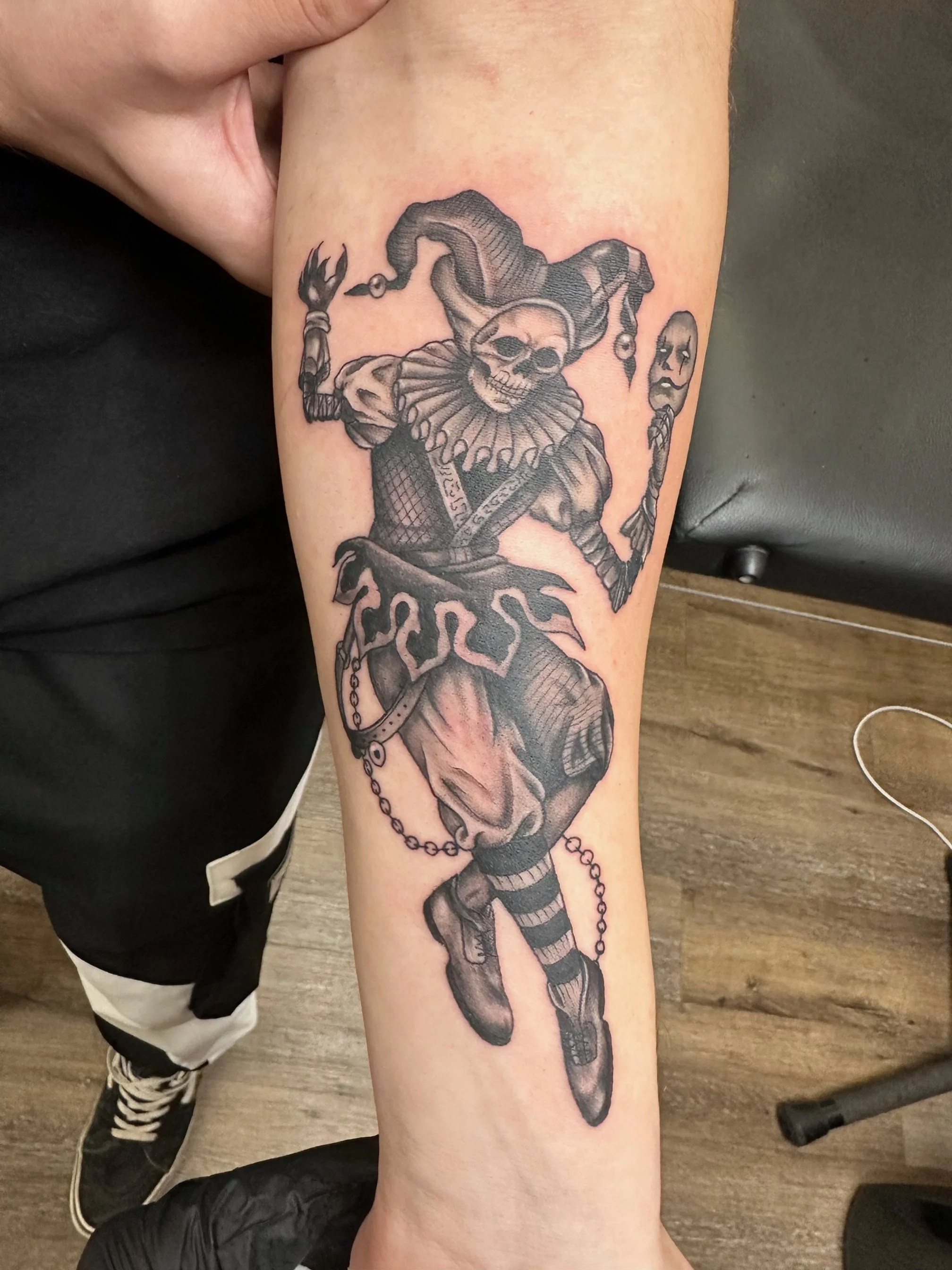 A darkly playful skeleton jester rendered in detailed black and grey shading. The piece balances macabre humor with intricate texture, creating a captivating gothic aesthetic.