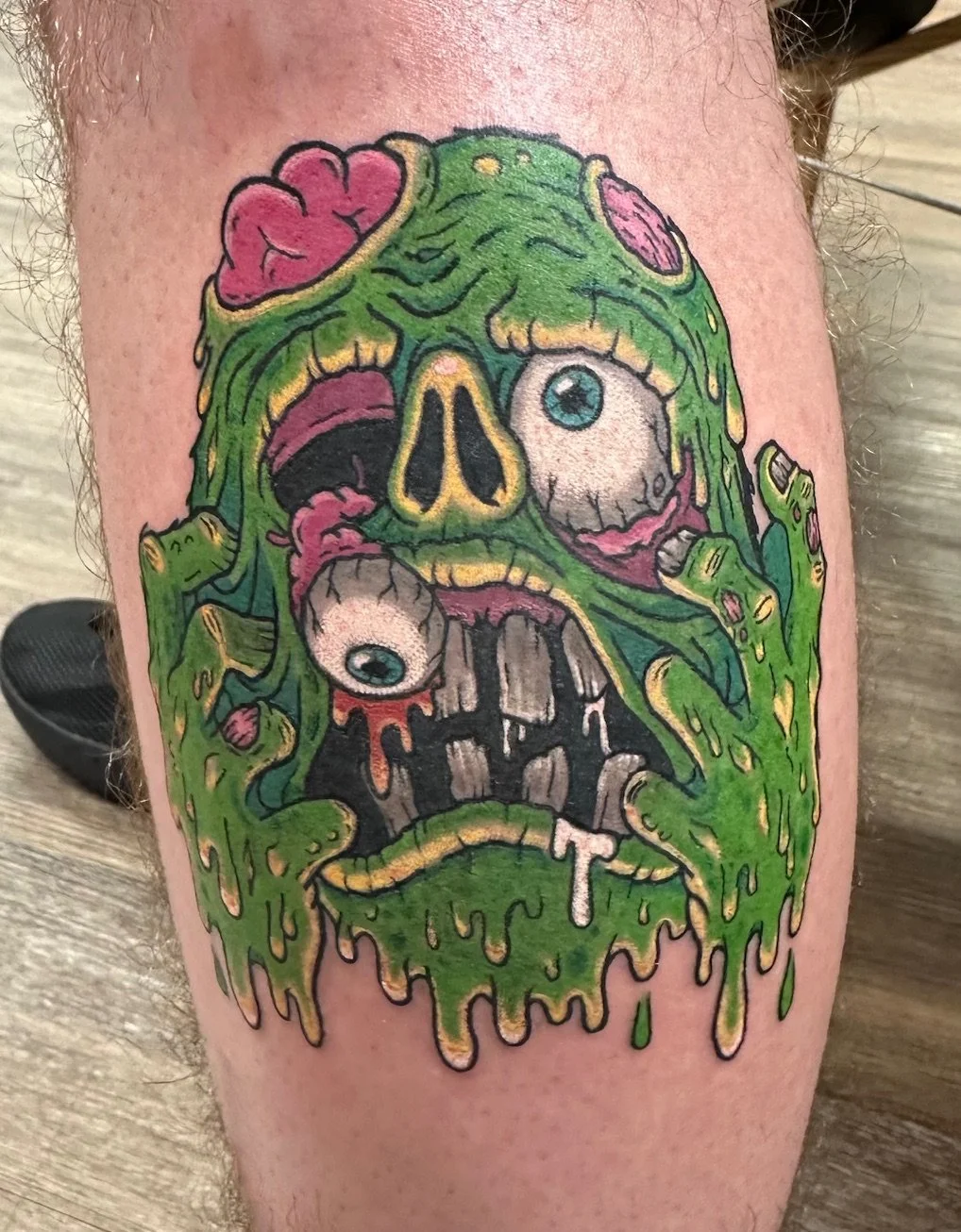 This bold, cartoon-horror tattoo showcases a grotesque melting zombie head in vibrant greens and pinks. The dripping textures and exaggerated features make it both eye-catching and playfully gruesome.