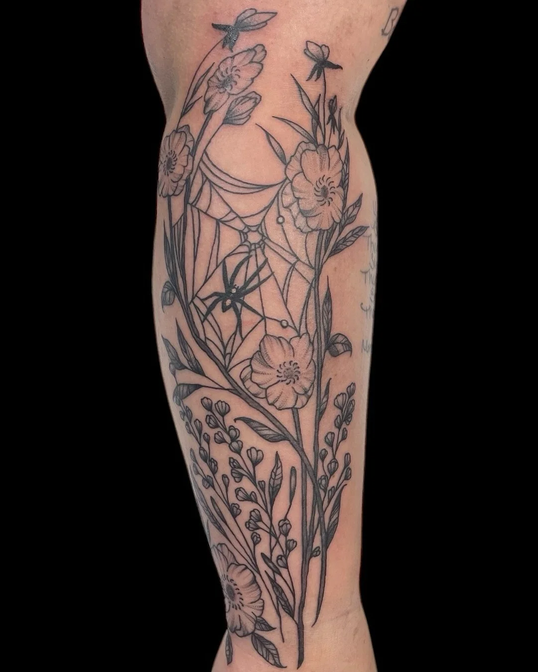An intricate blackwork design combining blooming wildflowers with a spider web at the center. The detailed linework and fine shading create a balance of natural grace and gothic undertone.