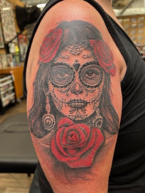 A detailed black and grey portrait with deep red roses, inspired by Día de los Muertos motifs. The shading and line detail add dimension and reverence to this striking piece.