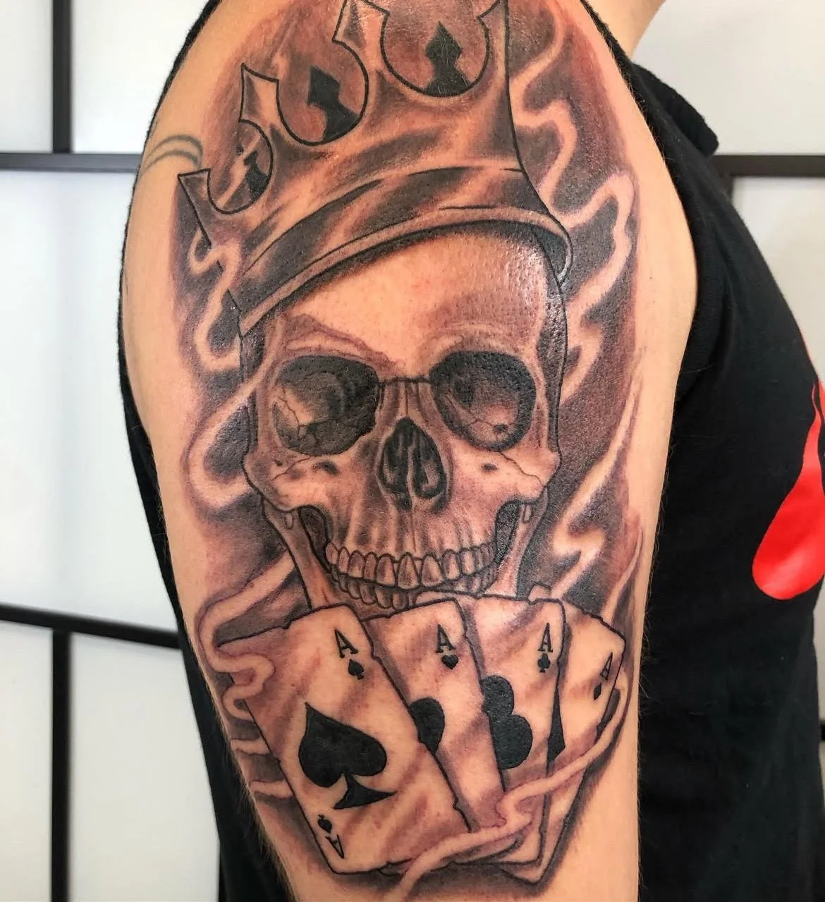 A menacing skull wearing a crown of flames and holding four aces. The bold black and grey shading and dramatic highlights give it a fierce, commanding presence.