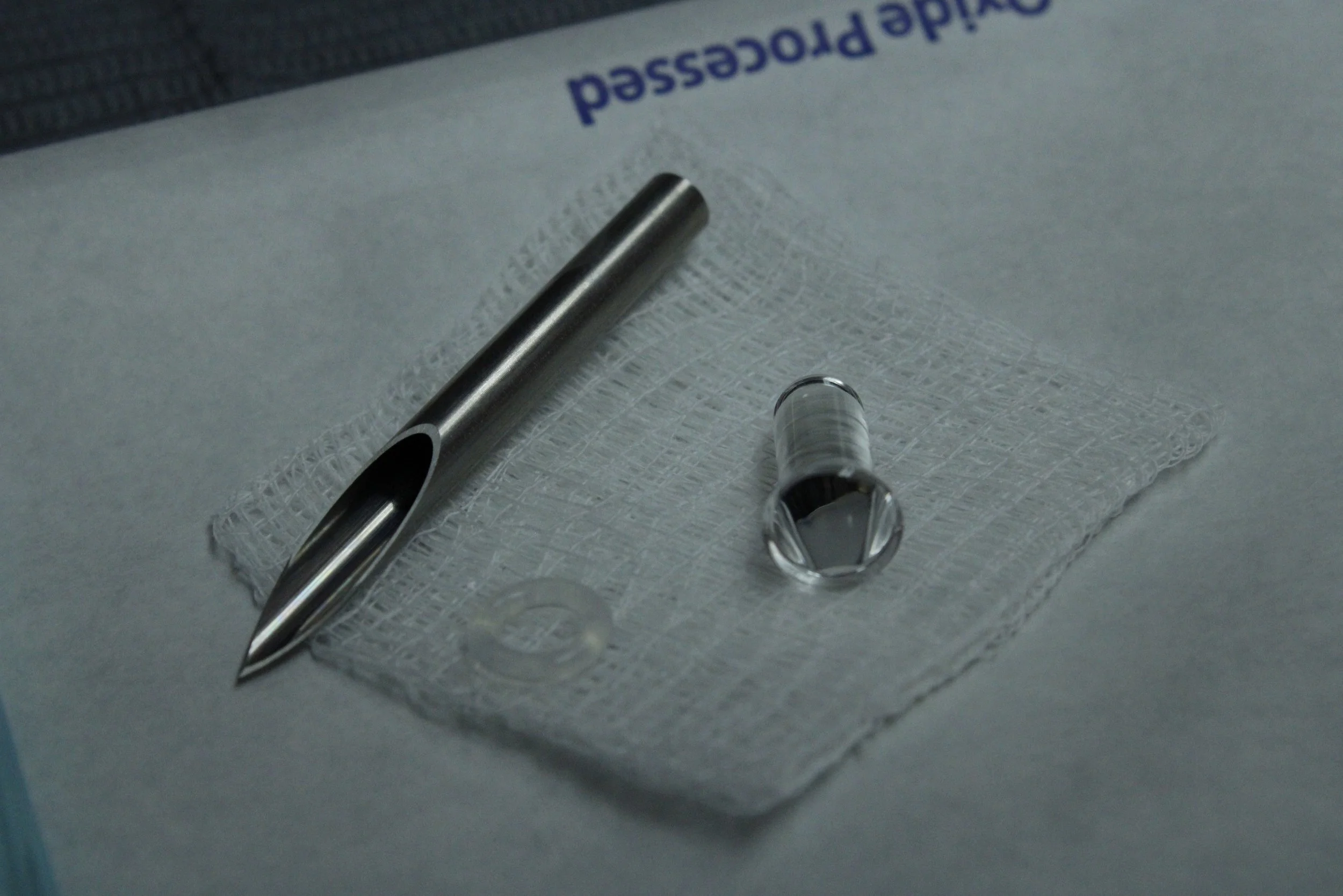 Medical tools including a metal needle, a glass vial with a dropper, and a small sterile gauze pad on a medical surface.