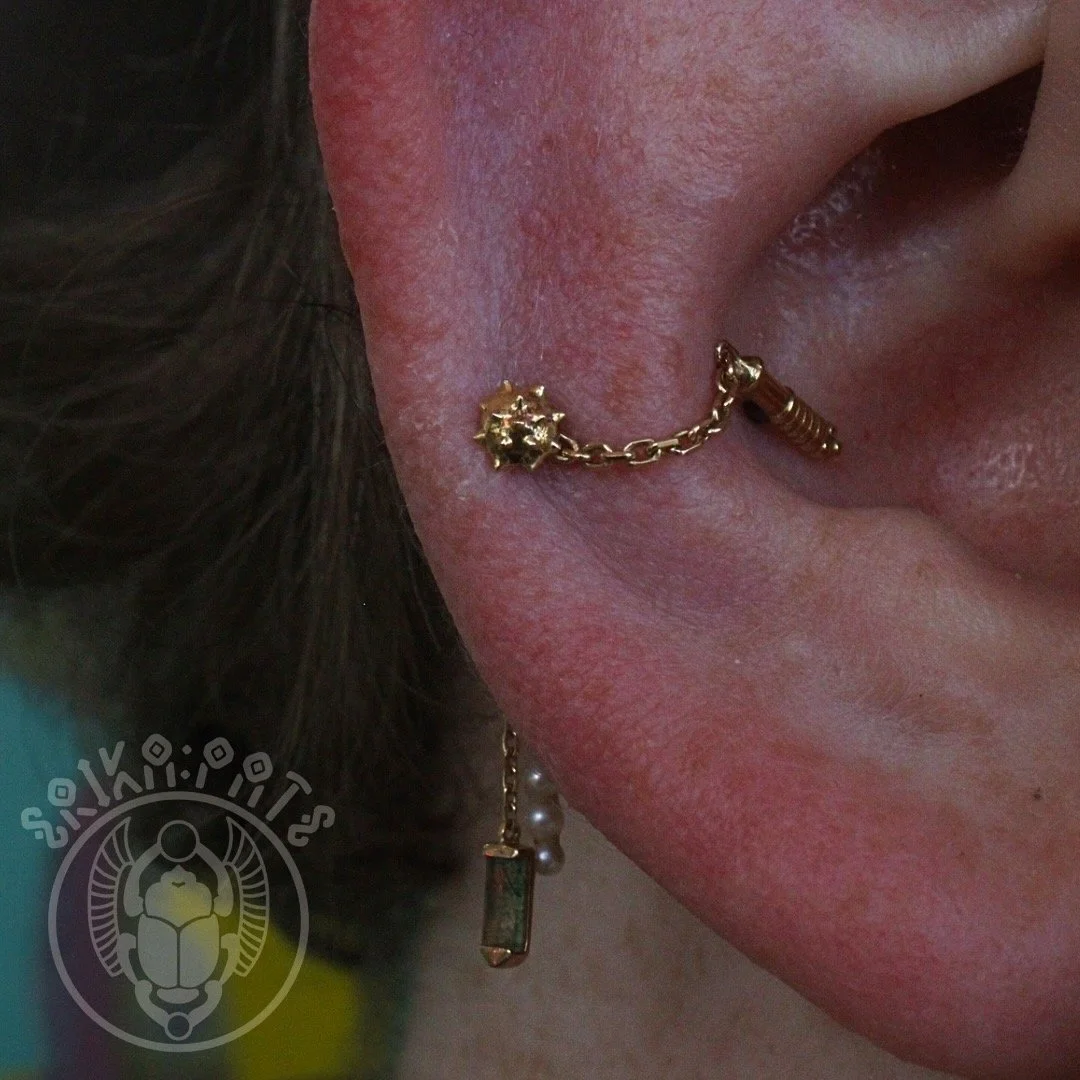An industrial piercing connects two cartilage points with a decorative gold chain, blending edge and sophistication. This bold style requires precise placement for comfort and long-term wear.