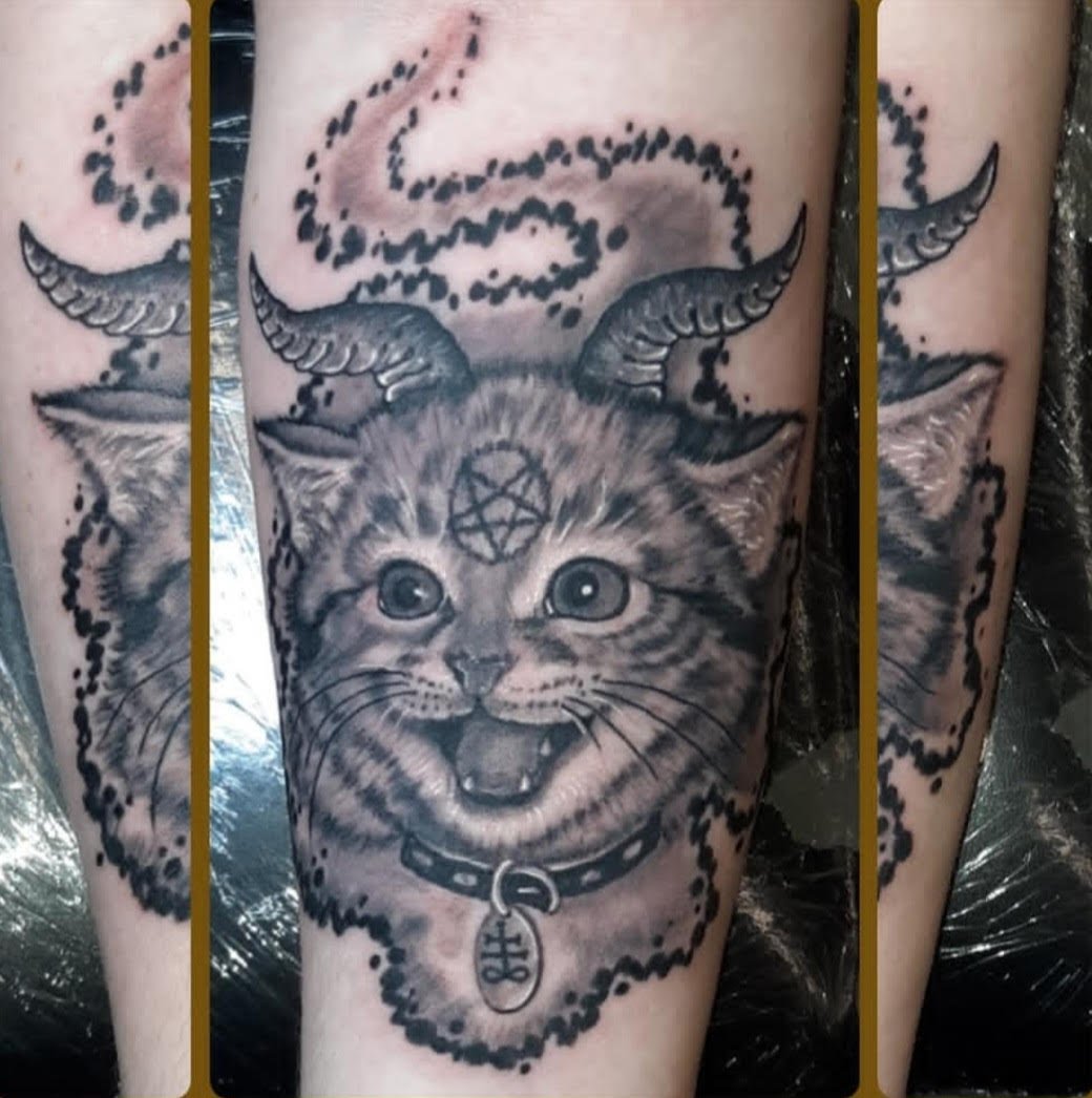 A highly detailed black-and-grey cat is depicted with horns, a pentagram symbol, and an ornate collar, blending realism with dark-fantasy elements. Dotwork shading adds texture while the mischievous expression gives the tattoo a playful yet sinister 