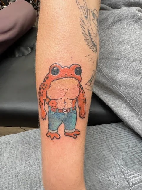 A humorous tattoo of a muscular frog standing upright in jeans with a confident stance. Its playful expression and cartoon realism make it both eye-catching and fun.