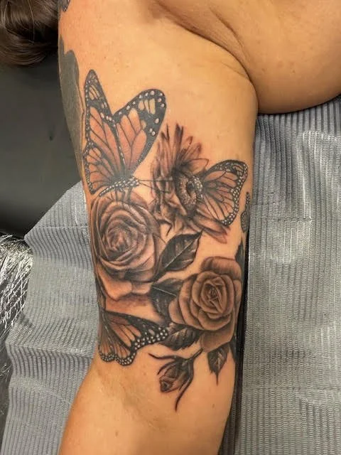 A black and grey floral sleeve featuring roses, sunflowers, and monarch butterflies. The delicate shading and natural flow combine realism with soft, organic beauty.