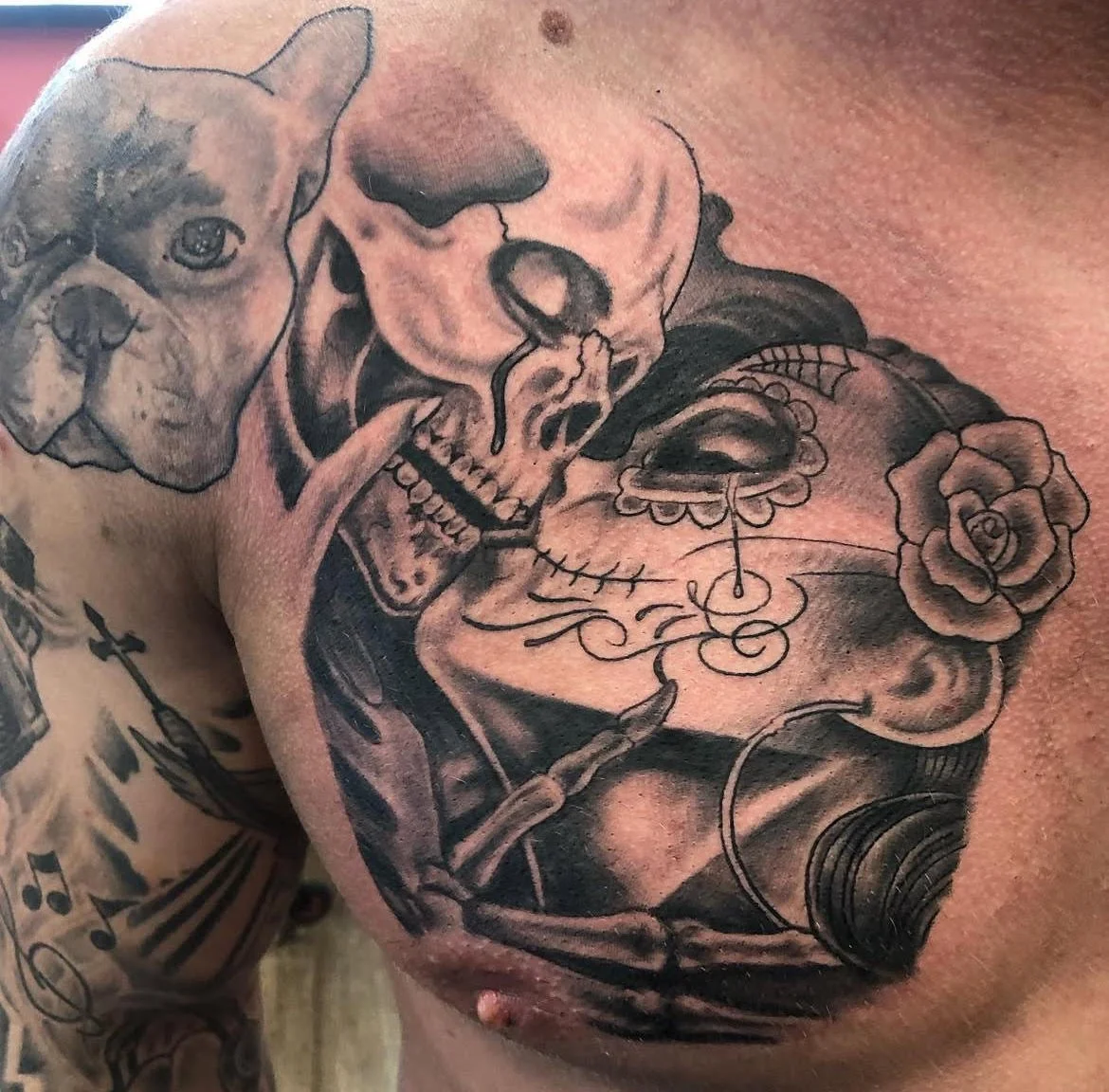 A dramatic black and grey chest piece featuring a skeletal figure kissing a woman in Día de los Muertos style makeup. The detailed shading and emotional imagery blend themes of love, life, and mortality.