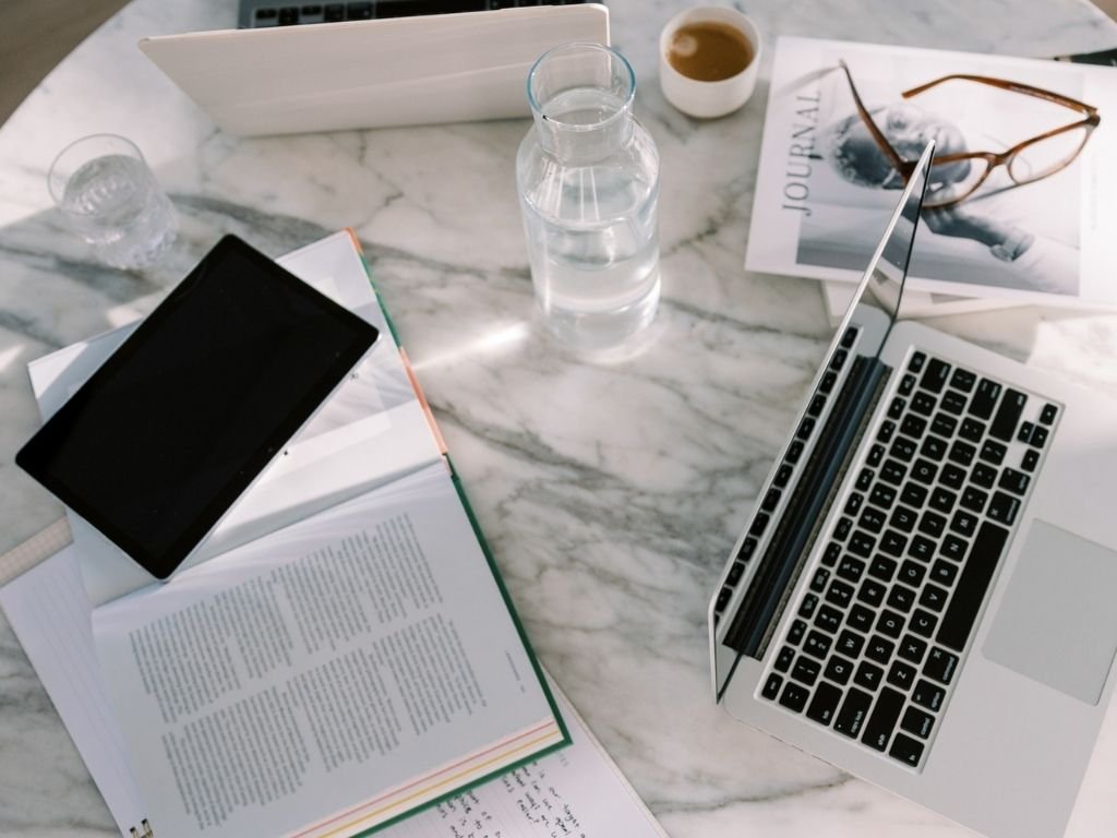 A marble desk with an open book, a black tablet, a glass of water, a glass water bottle, a laptop, a small cup of coffee, glasses, and a journal on it.