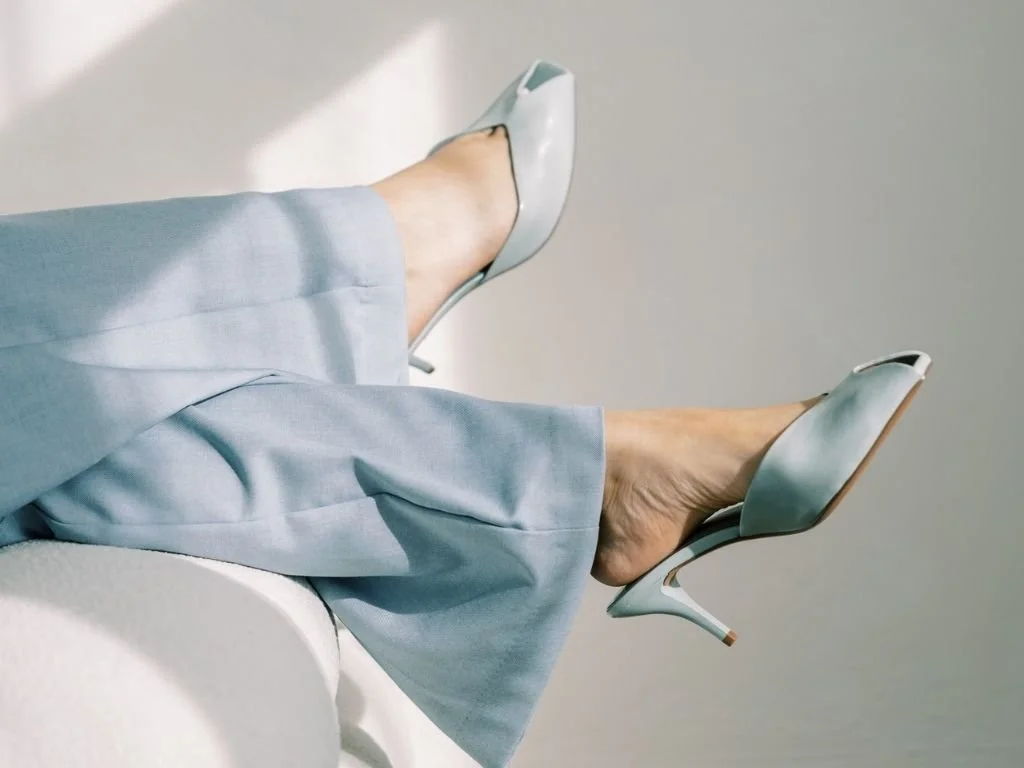 A person wearing light blue pants and high heel shoes sitting on a white surface with legs crossed